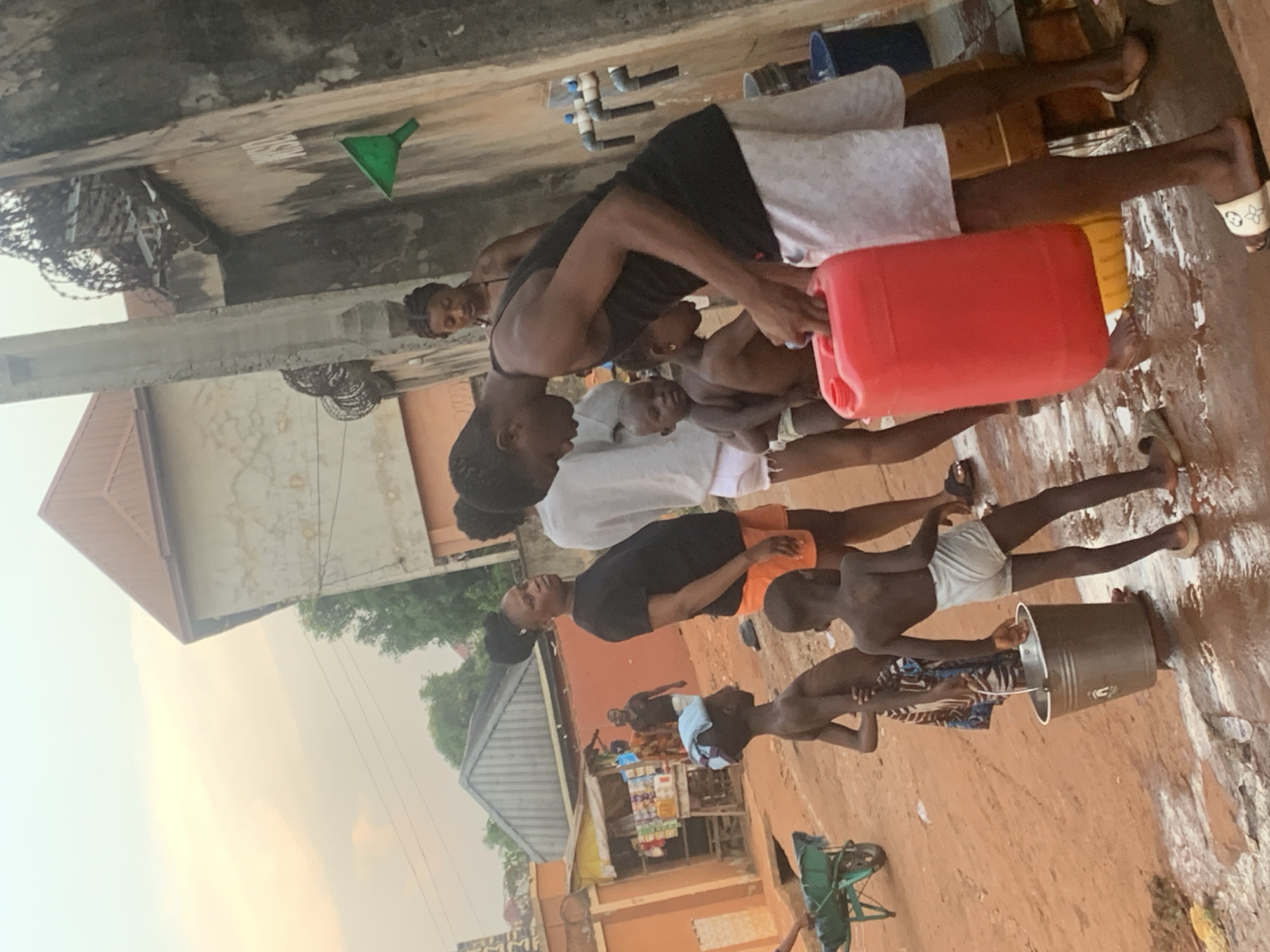 In Ukwu-Abwa, Nigeria, a group of people stand outside on a red dirt ground. A woman wearing a black top and white shorts pours liquid from a red container for a group of children. The children stand in a line, some holding containers such as a metal bucket. Other people are visible in the background near a building that appears to be a store or stall. Buildings and trees are also visible in the background. The sky is overcast.