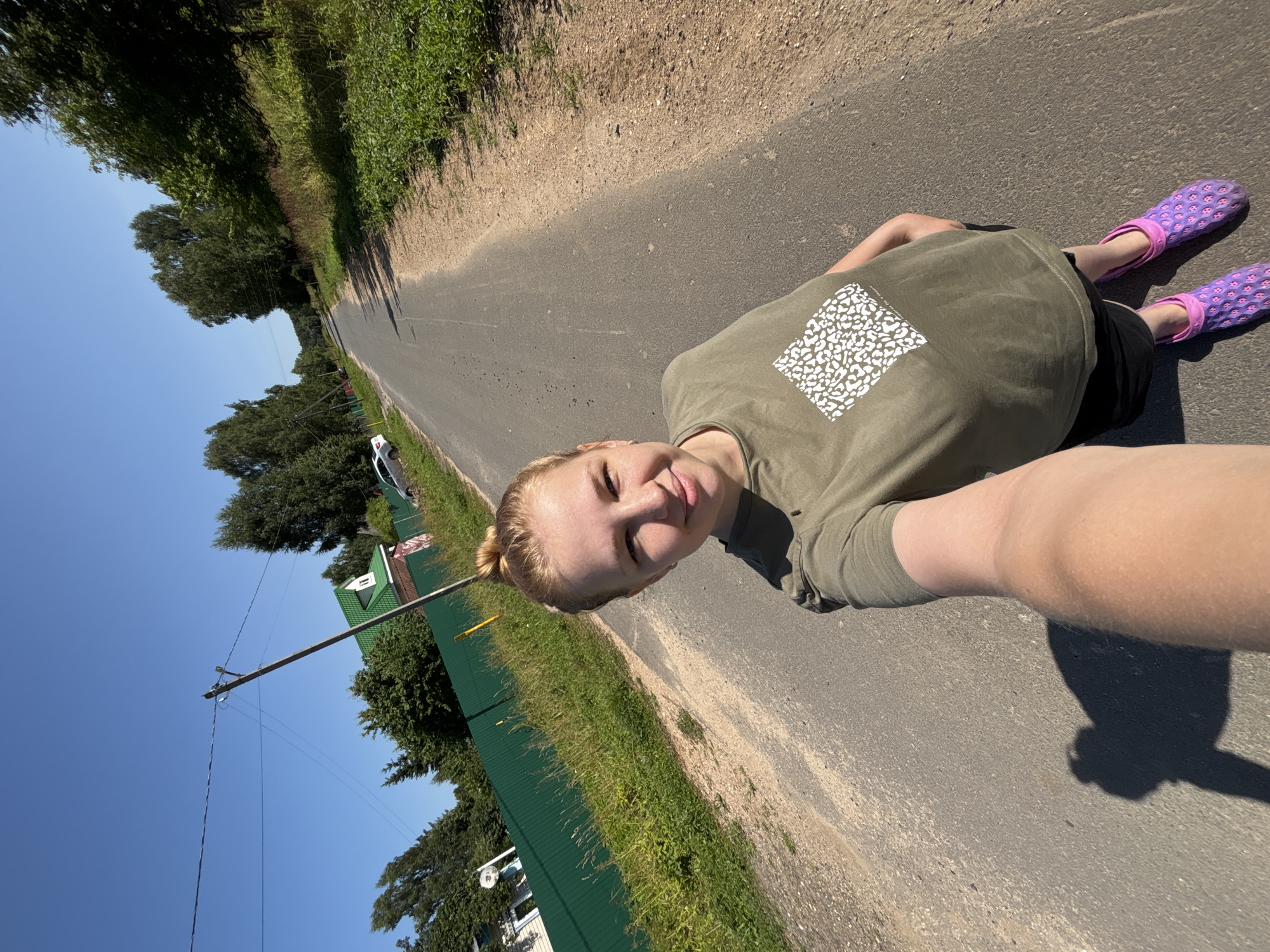 A young woman is taking a selfie while standing on a paved road in the village of Dunilovo, Russia. She is wearing a green t-shirt and black shorts, and bright pink clogs. The sun is shining brightly, suggesting it is daytime and the weather is warm. The road is bordered by a grassy verge and a green fence, with trees and houses visible in the background. A utility pole with wires stands to the left of the woman, reaching towards the clear blue sky. The woman is smiling at the camera, appearing happy and relaxed. The image is oriented sideways.