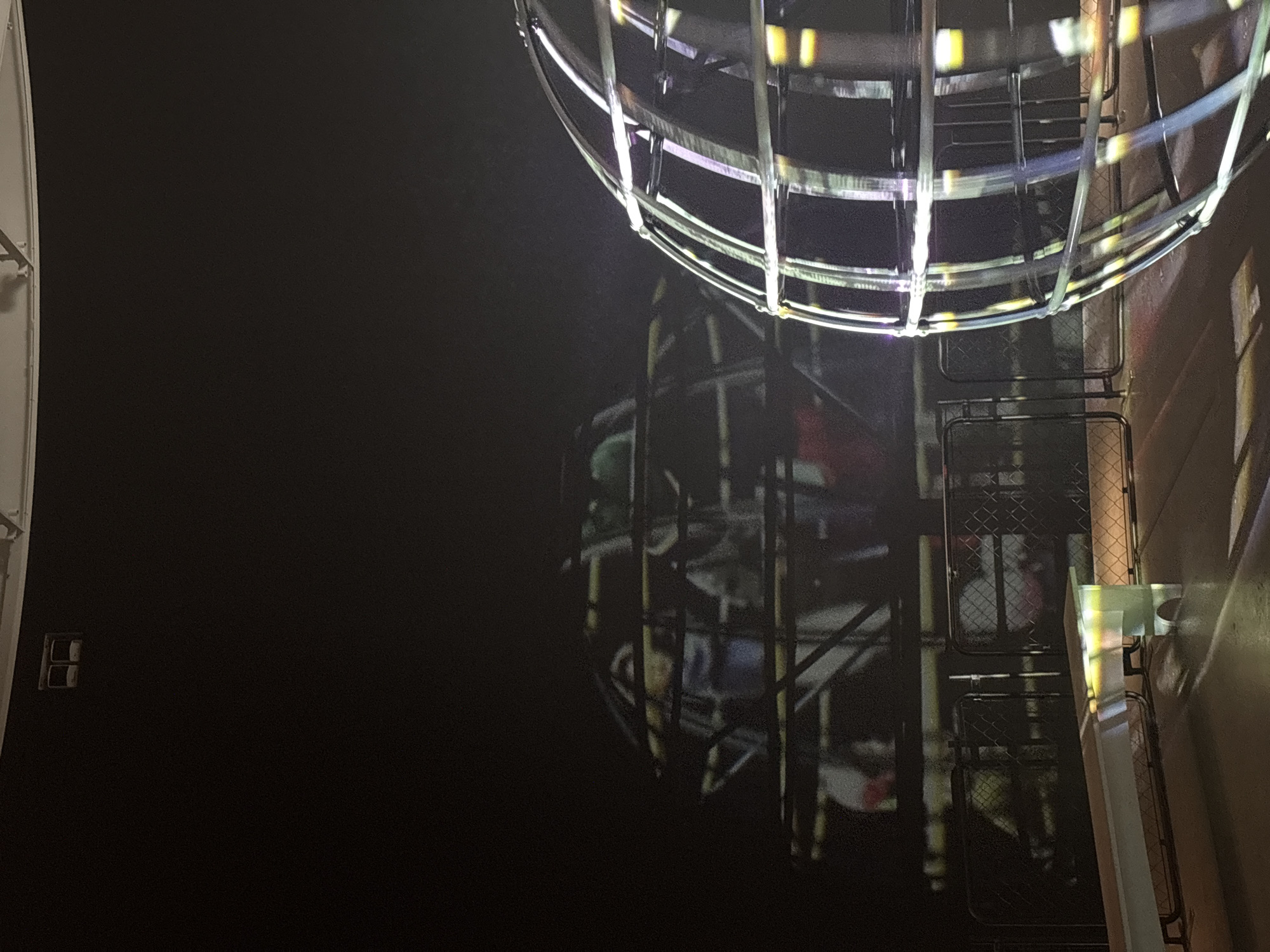 This media file captures a dark, indoor scene, strongly suggesting an art museum or contemporary exhibition space in Kanazawa, Japan. The focal point is a large, luminous spherical structure, positioned in the upper right. This sphere is constructed from bright, metallic bars or tubes that glow with white and faint colorful light, possibly from internal illumination or reflected sources, resembling a modern art installation.
Below and to the left of the glowing sphere, a faint, ethereal image is projected or reflected onto the dark wall. This ghostly projection depicts indistinct figures, possibly people, situated within a cage-like or multi-tiered structure, with subtle hints of color, including red. This element suggests an interactive light projection or a reflective surface within the exhibit.
Along the right edge of the frame, a light-colored vertical structure, likely a wall or pillar, features a translucent panel that emits soft, rainbow-colored light, adding another layer of illumination to the scene. Below this, a dark metal railing or part of an exhibit barrier is visible. On the far left, a small, dark rectangular object, possibly an electrical outlet or switch, is present on the dark wall.
The lighting is predominantly dim, typical for showcasing light installations, creating a mysterious and immersive atmosphere. No discernible text is visible in the image.