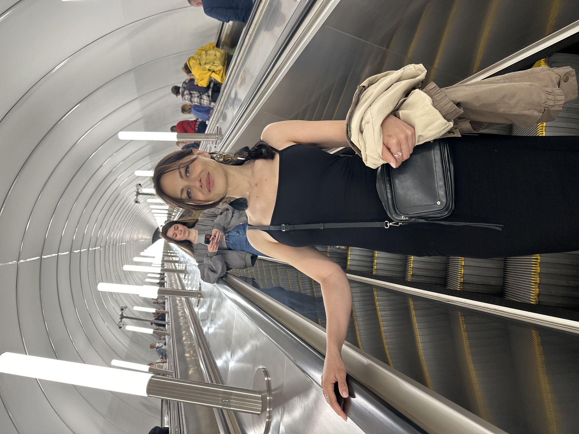 The image depicts a woman standing on an escalator in the Saint Petersburg Metro. The escalator is wide with silver handrails and yellow-lined steps, ascending into a brightly lit tunnel with a curved, white ceiling. Tall, cylindrical light fixtures line the tunnel's sides. The woman is wearing a black, spaghetti-strap dress and a light-colored jacket draped over her left arm. She has dark hair and is holding a black purse with a strap across her body. Her left hand rests on the escalator handrail, and she is wearing a ring with a light-pink stone on her left ring finger. In the background, other passengers are visible on the escalator, some looking at their phones, and others facing forward. The environment suggests a modern and well-maintained underground public transport system, characteristic of the Saint Petersburg Metro.