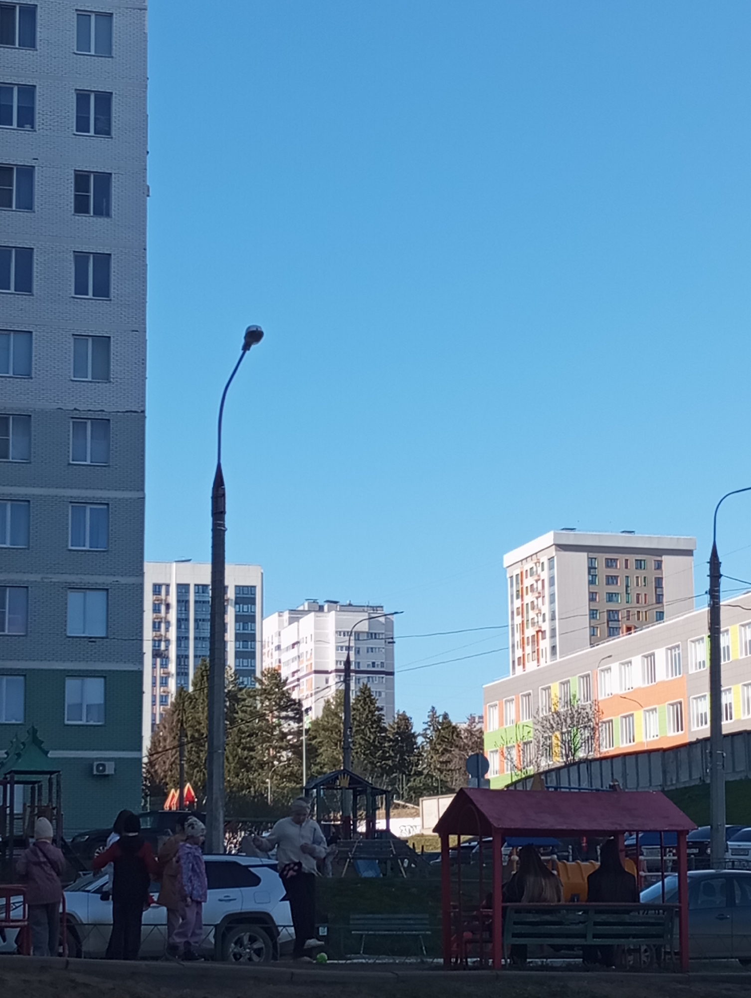 The image shows a sunny day in what appears to be a residential area in Izhevsk, Russia, with several multi-story apartment buildings visible in the background. A group of people, including several children, are standing or interacting near a parked white SUV. There is a playground area with a small red-roofed structure where people are seated. A lamppost stands prominently in the foreground. The buildings are typical of urban housing, with repeated window patterns. The environment suggests a common outdoor space adjacent to residential buildings. The weather is clear with a bright blue sky.