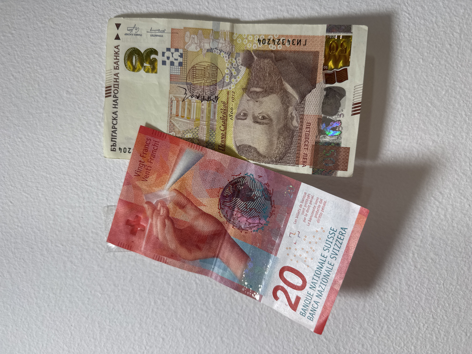 A top-down, brightly lit photograph displays two banknotes, a Bulgarian 50 Lev and a Swiss 20 Franc, laid on a light-colored, textured surface. The Bulgarian 50 Lev note is positioned horizontally at the top, featuring a prominent portrait of Pencho Slaveykov with his birth and death years (1866-1912), alongside an architectural illustration. Text includes "БЪЛГАРСКА НАРОДНА БАНКА" (Bulgarian National Bank), "ПЕТДЕСЕТ ЛЕВА" (Fifty Leva), the numeral "50", signatures for "УПРАВИТЕЛ" (Governor) and "ГЛАВЕН КАСИЕР" (Chief Cashier), and the serial number "ГИЗ4324204". Part of the year "204" is also visible. This note is predominantly beige, yellow, and brown, with iridescent security features.
Below it, angled diagonally, is the red and orange Swiss 20 Franc note. It depicts a human hand in a gesturing pose and a stylized, patterned globe. Visible text includes the numeral "20", "Vingt Francs", "Venti Franchi", "BANQUE NATIONALE SUISSE", "BANCA NAZIONALE SVIZZERA", and a security warning in French and Italian: "Les billets de banque sont protégés par le droit pénal. Le banconote sono protette dal diritto penale." A white cross, part of the Swiss emblem, is also visible. The background is a uniform, subtly textured white or off-white material, providing a neutral backdrop for the currency.