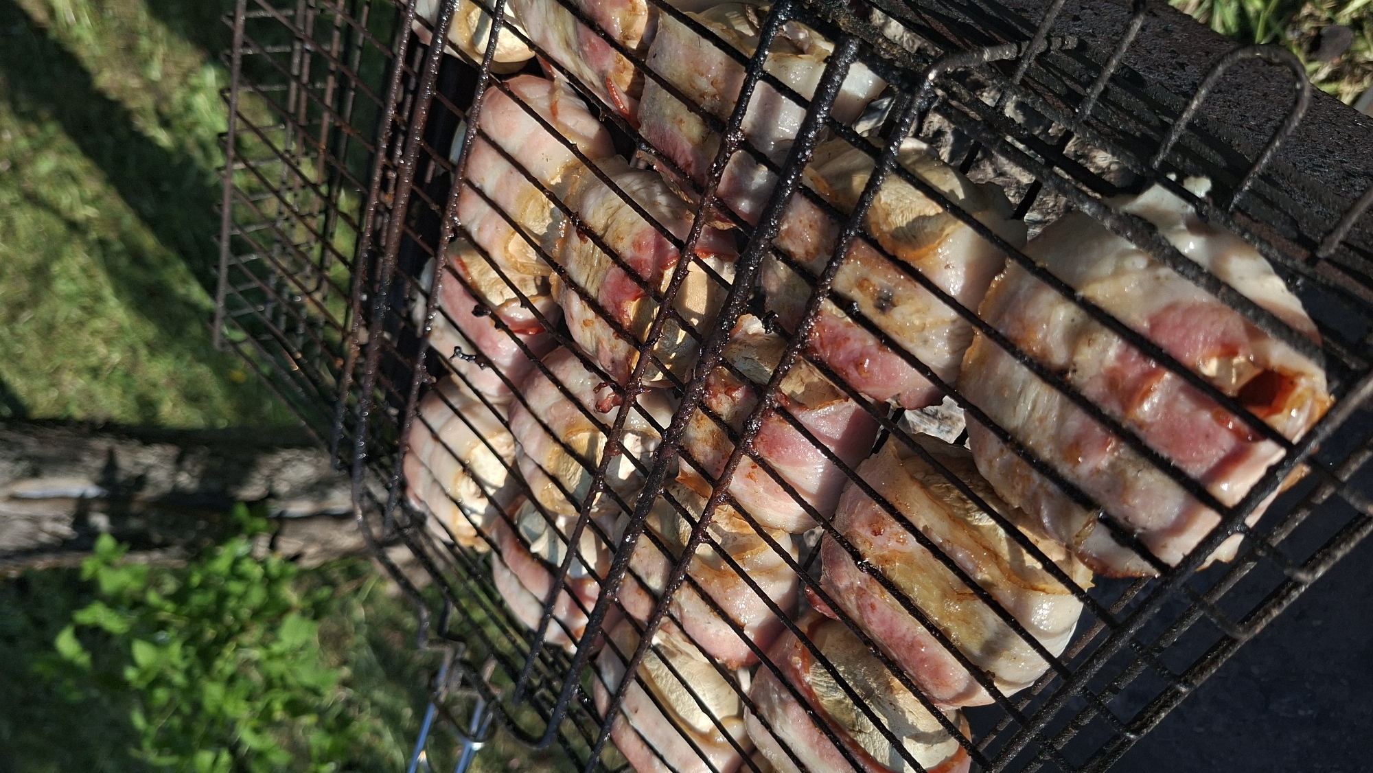 The image depicts a metal grill filled with food items. Specifically, it appears to be mushrooms wrapped in bacon, suggesting they are being cooked or grilled. The grill is made of a dark, possibly rusty metal, with a grid pattern that holds the food in place. The mushrooms are circular, light-colored, and wrapped with strips of bacon, which are pink and white with some browning from cooking. The background shows a glimpse of a green grassy area, indicating an outdoor setting. The image appears to have been taken in Сваткаўскі сельскі Савет, Belarus.