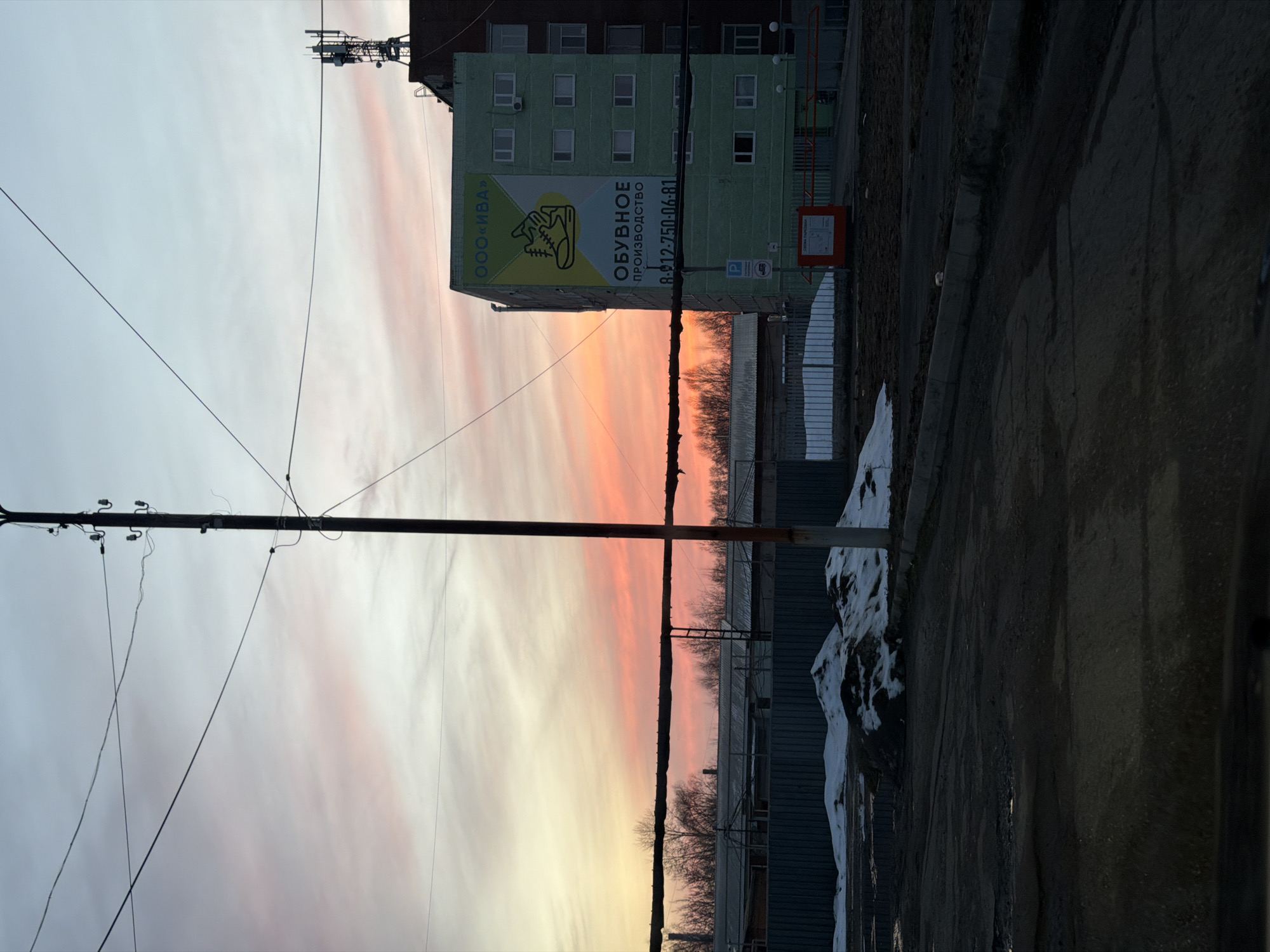 The image shows an urban scene in Sarapul, Russia at sunset. A telephone pole with several wires is visible on the left side of the frame. A multi-story, light green building is present, with a large advertisement for "ОБУВНОЕ ПРОИЗВОДСТВО" (Shoe Production) and the phone number 8-912-750-06-81 on its side. The sky behind the building has a vibrant orange and pink hue, indicating the sunset. A chain link fence and a small pile of snow are located on the right side of the image. The lower part of the frame contains a dark concrete wall, with a blurry image of a dark vehicle.