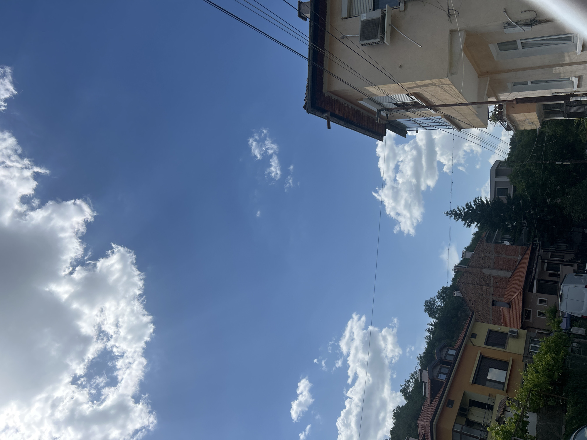The image depicts a view looking up at a clear blue sky with scattered white clouds, framed by buildings and power lines. The buildings, seemingly located in Unknown, Unknown, are diverse in style, with varying roof colors and architectural details. The environment suggests an urban or suburban setting with greenery visible among the buildings. No people are visible.