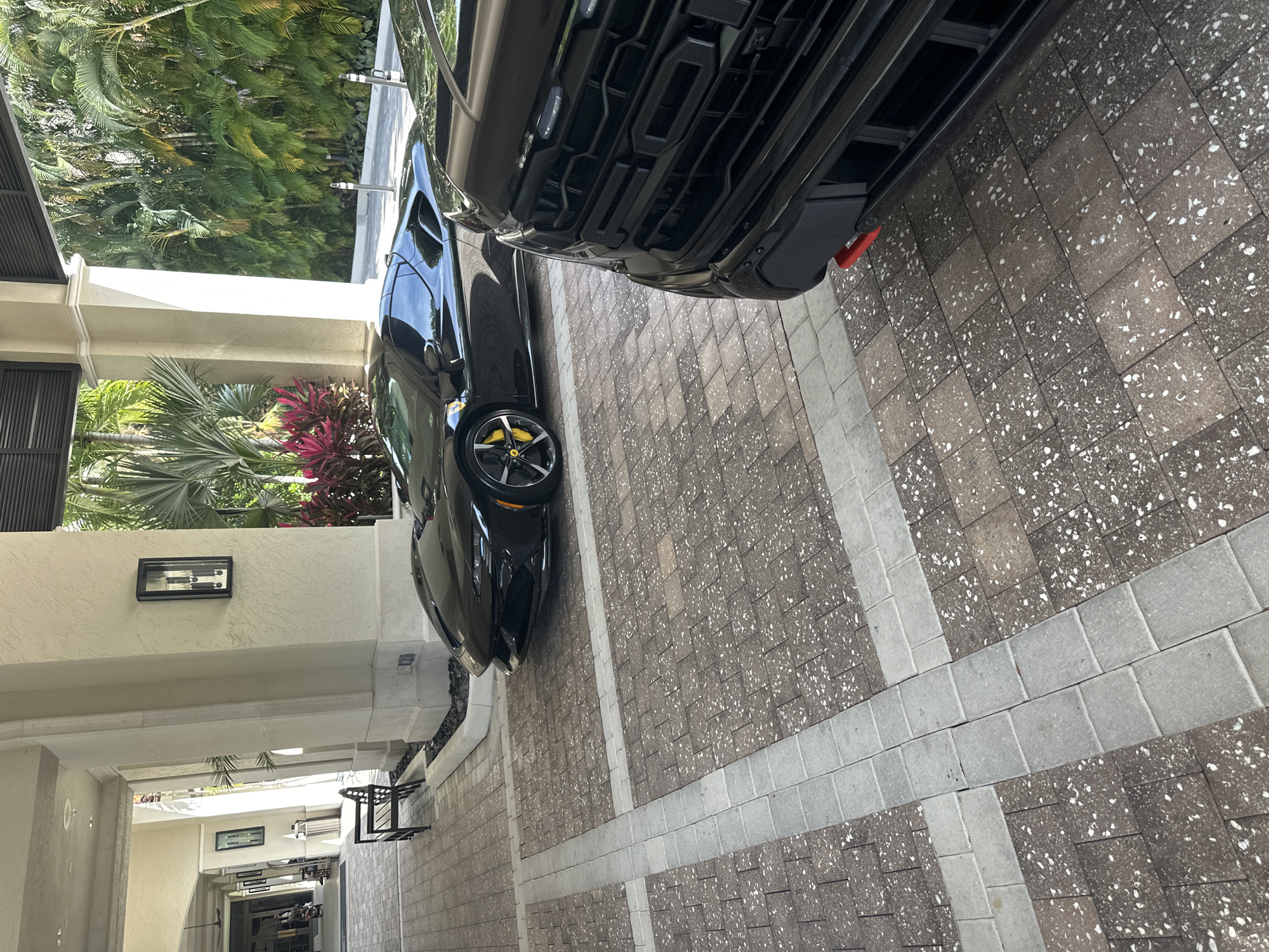The image captures a view of a driveway with two vehicles parked. The driveway is constructed of rectangular pavers arranged in a grid-like pattern, with alternating sections of lighter and darker tones. On the left of the frame, a portion of a building is visible, featuring an exterior wall with a light fixture and a covered walkway with additional architectural details.
A black sports car with distinctive yellow brake calipers is parked on the left side of the frame, closer to the building. To the right of the sports car, the front end of a larger, dark-colored vehicle is visible. The scene appears to be outdoors.
Based on the available details, it can be inferred that the location is within a residential area in the city of Aventura, United States of America.