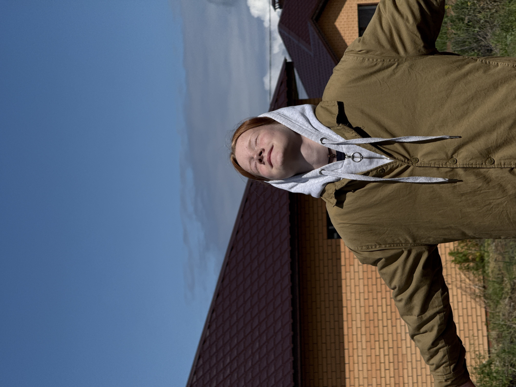 A reddish-haired young adult with freckles is standing in front of a brick and red-tiled house, wearing a tan jacket over a gray hooded sweatshirt. The sky is blue with some clouds. The house appears to be a brick and tile building in Chaikovsky, Russia.