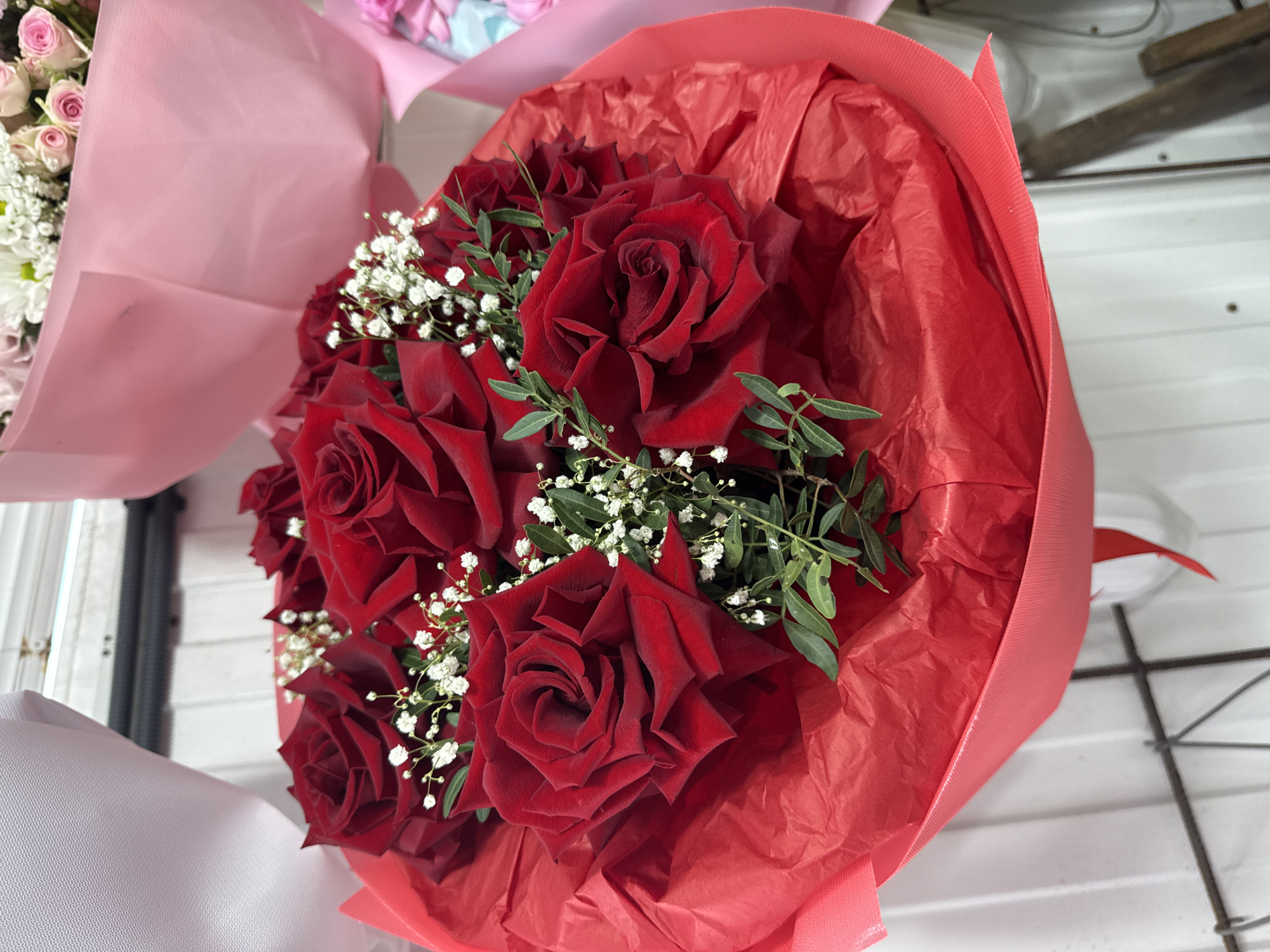 A bouquet of red roses is displayed in front of a white corrugated wall. The roses are arranged in a circular fashion and are complemented by small white flowers and green foliage. The bouquet is wrapped in red paper, and a pink ribbon is visible at the bottom of the image. The overall lighting suggests an indoor setting.