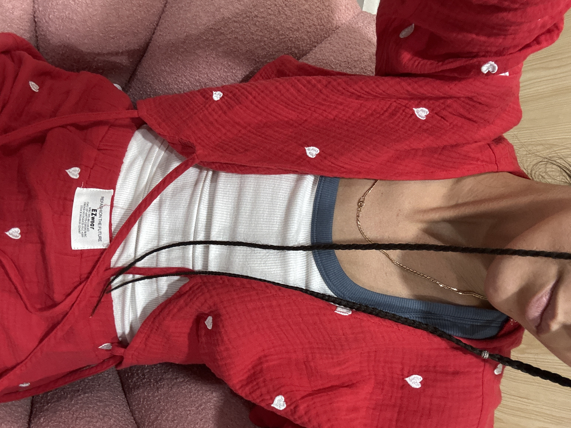 The image shows a close-up of a person wearing a red cropped jacket with white heart patterns over a white ribbed tank top and red shorts. The person has dark braided hair with beads. A gold chain necklace is visible around the neck. The clothing is set against a blurred background that appears to be a wooden wall. A pink textured surface is visible on the left and right sides of the frame, suggesting the person is seated. A white tag with black text, reading "REFASHION THE FUTURE" and "EZwear" is visible on the shorts. The image does not provide any specific information or context related to Nam Hoa Lư Ward, Vietnam.