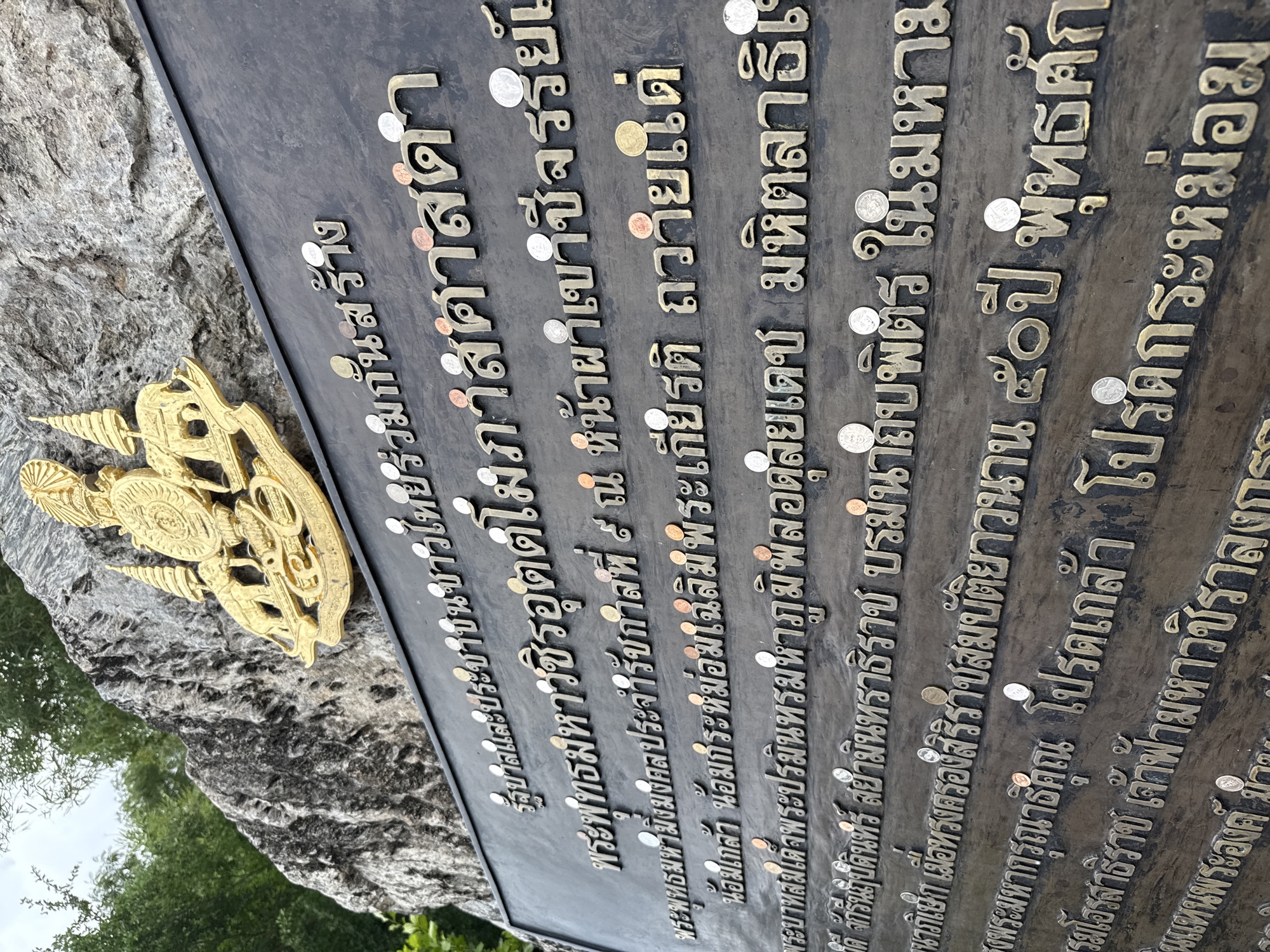The image shows a close-up of a plaque mounted on a rock face, likely outdoors, with trees in the background. The plaque is inscribed with Thai text in a raised, metallic font, and several coins are affixed to its surface. At the top of the plaque is a golden emblem featuring elephants and a spire. The plaque appears to be a commemorative or informational marker. Based on the context provided by the prompt, this scene is located in Na Chom Thian, Thailand.