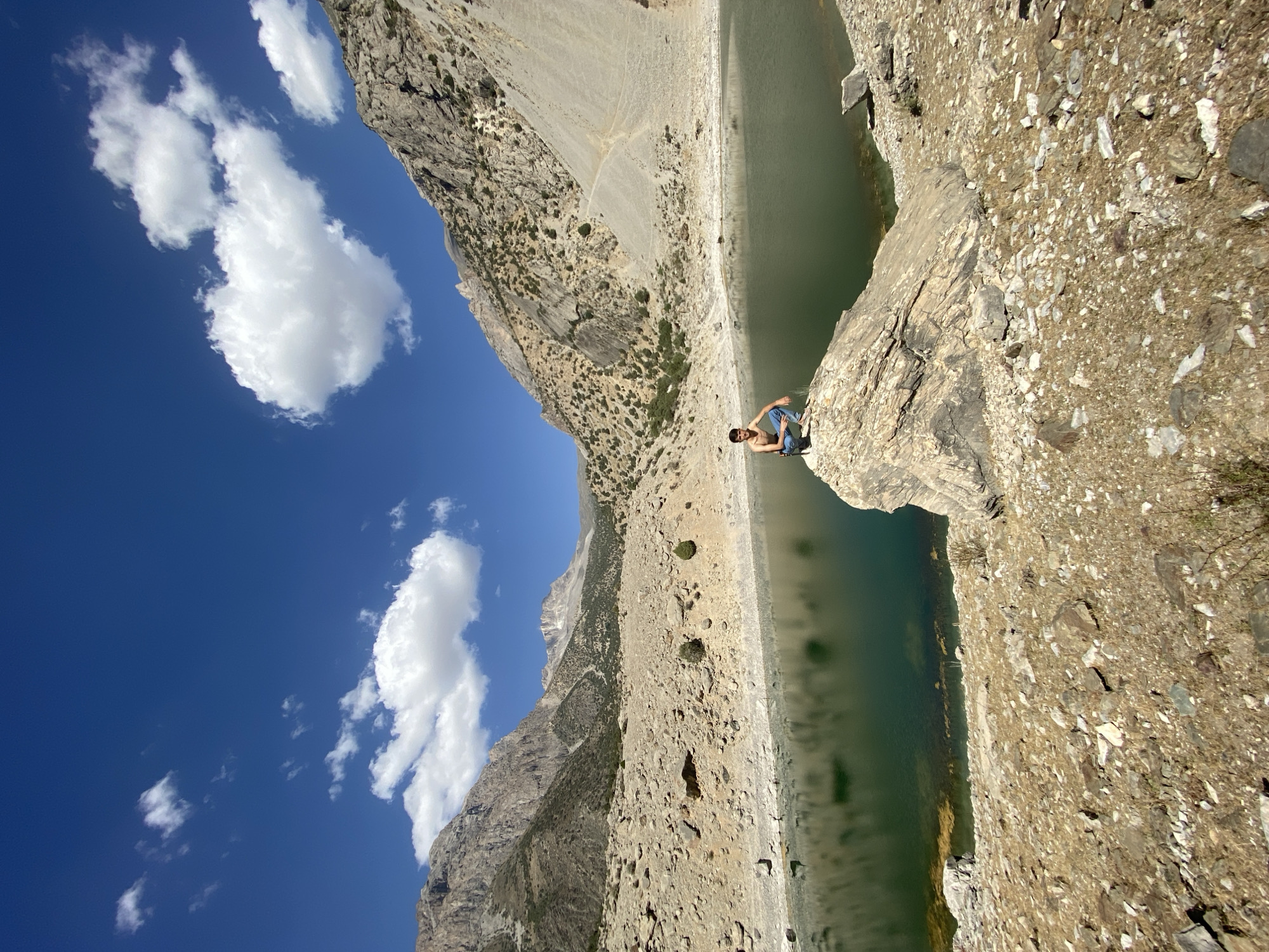 A young man, shirtless and wearing blue jeans, is seated on a large, weathered rock. He is looking towards the camera. The rock he is sitting on is in the foreground, close to a body of dark, calm water. Beyond the water, the landscape consists of arid, rocky hills and mountains. The terrain is sparsely vegetated with small shrubs and grasses. In the background, large, imposing mountains rise towards a bright blue sky dotted with fluffy white clouds. The sun appears to be high in the sky, casting shadows and illuminating the scene. The location is identified as Ghazza, Tajikistan.