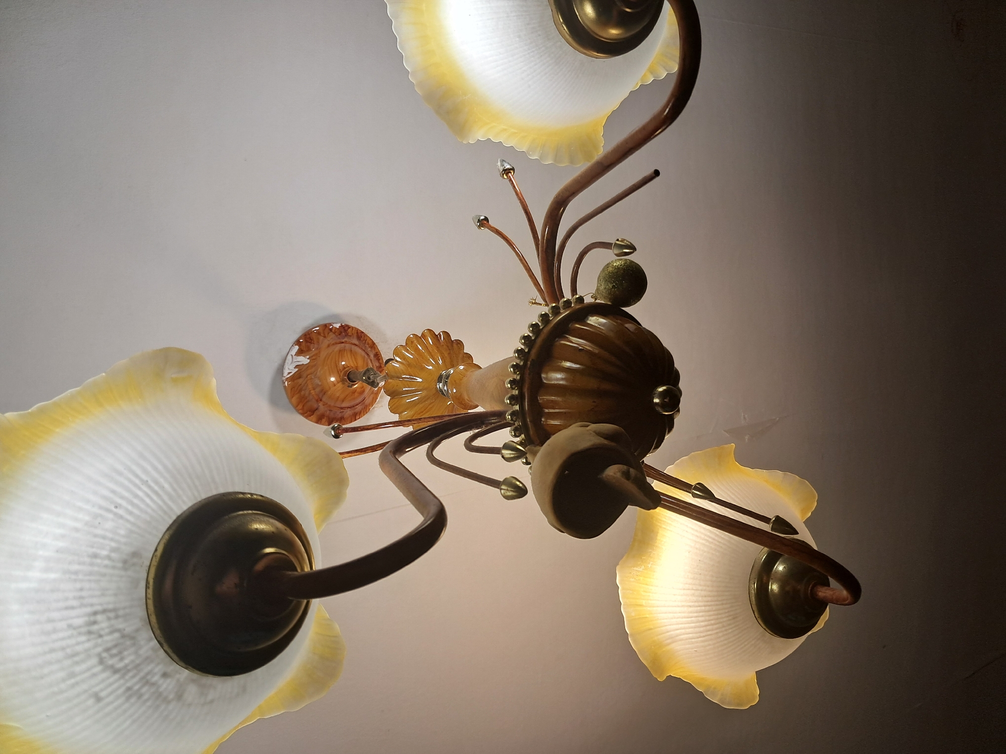 An ornate, multi-tiered chandelier hangs from a white ceiling. The chandelier features several frosted glass shades with yellow edges, shaped like open flowers or fans. The shades are supported by dark brown, curved arms that connect to a central wooden body. This central body is decorated with ribbed detailing and brass accents, including a beaded band and a finial at the top. Several thin, copper-colored arms extend from the central body, some ending in decorative finials, and one holding a small, green, spherical ornament. The lighting is dim, with warm light emanating from the shades. The background is a plain, white ceiling.