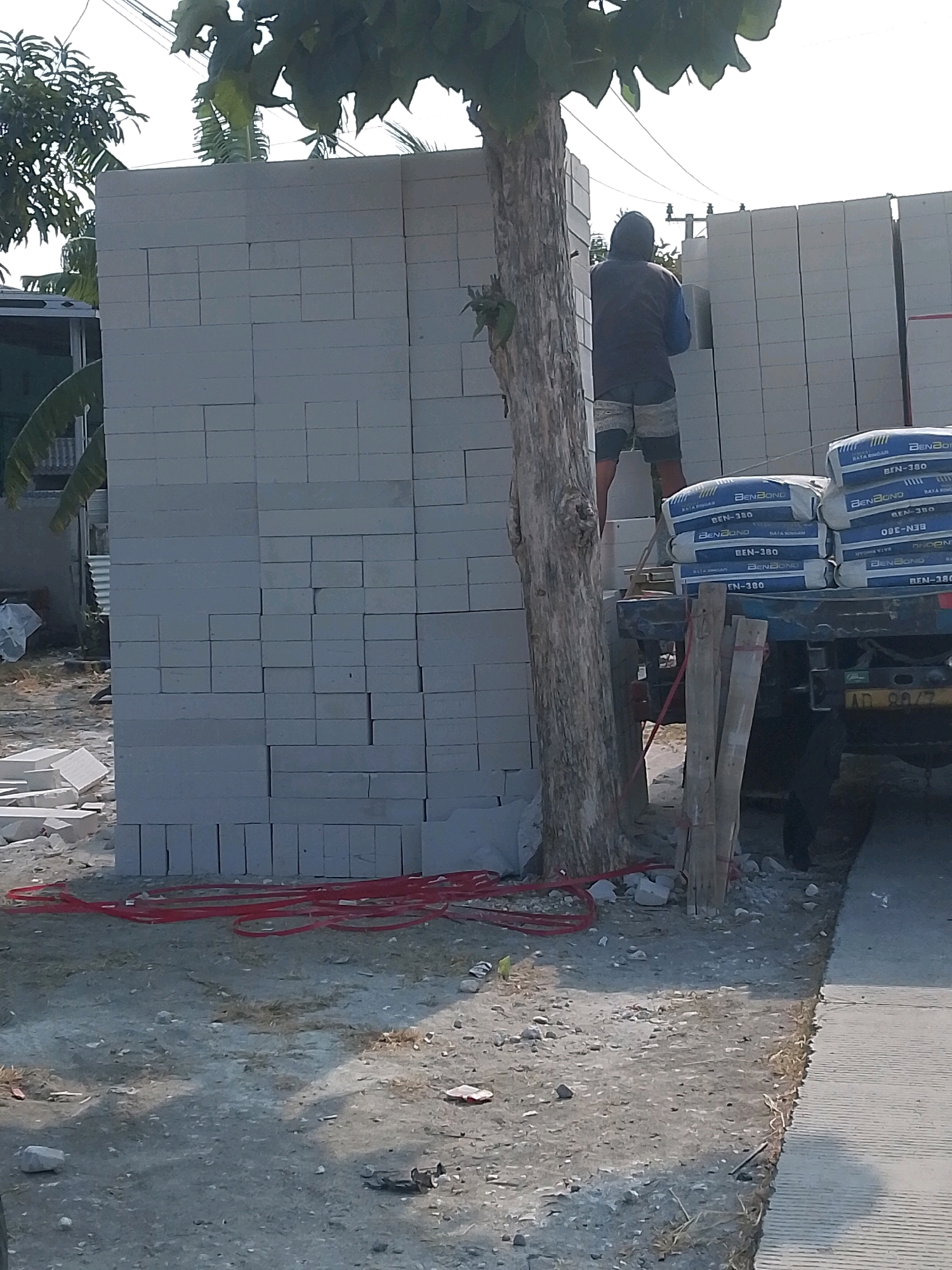 A male construction worker, wearing a dark hooded vest over a blue long-sleeved shirt and patterned shorts, stands on a ladder against a tall stack of light grey concrete blocks, reaching up to place another block. He is positioned in front of a large wall constructed from similar blocks. To the right of the wall, several large white sacks, labeled with blue lettering that includes "BenBond" and "BEN-380," are stacked on the back of a dark-colored truck. The truck has a yellow license plate with the characters "AD 9247". A weathered tree trunk with some foliage stands prominently in front of the block wall and slightly to the left of the worker. Red electrical wiring is loosely coiled on the dusty ground in the foreground, near the base of the tree and extending towards the truck. In the background, a building with a corrugated metal roof and palm fronds are visible. The scene appears to be an outdoor construction site with abundant sunlight and a clear sky.