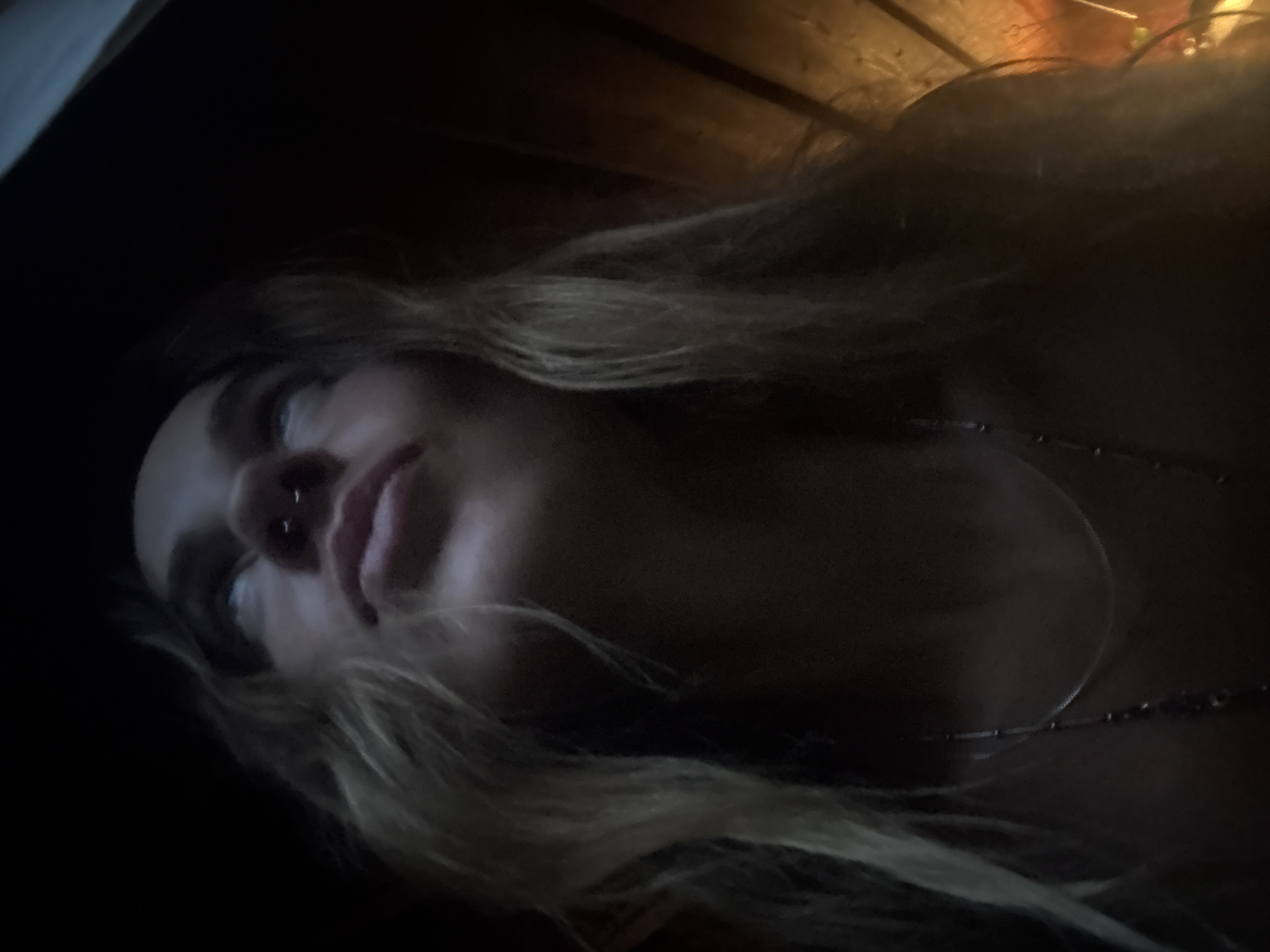 A low-angle, close-up, indoor shot shows a young person with blonde, wavy hair looking upwards. Their face is illuminated by an unseen light source, creating dramatic shadows. The person has a nose ring and is wearing a necklace. The background is dark and appears to be made of wood. The image is blurry and grainy, suggesting it may have been taken in low light conditions. The location is unknown.