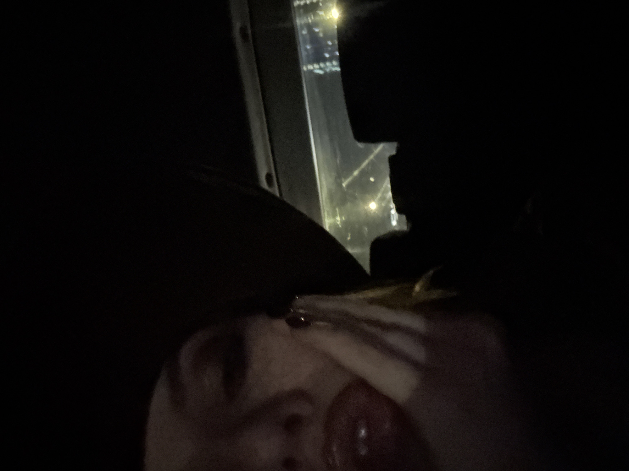 The image shows a close-up of a person's face, with a portion of a dark hat visible on their head. Their lips are painted red and slightly open. Their right hand is raised towards their face, with painted nails. The background is dark, with the reflection of city lights visible through a window, suggesting the subject is likely in a vehicle at night. The city lights appear to be from Sofia, Bulgaria, indicated by the presence of the National Palace of Culture (NDK) in the background, a prominent landmark known for its illuminated facade at night. The person's expression and pose are suggestive of a selfie taken in a moving vehicle or a dimly lit environment.