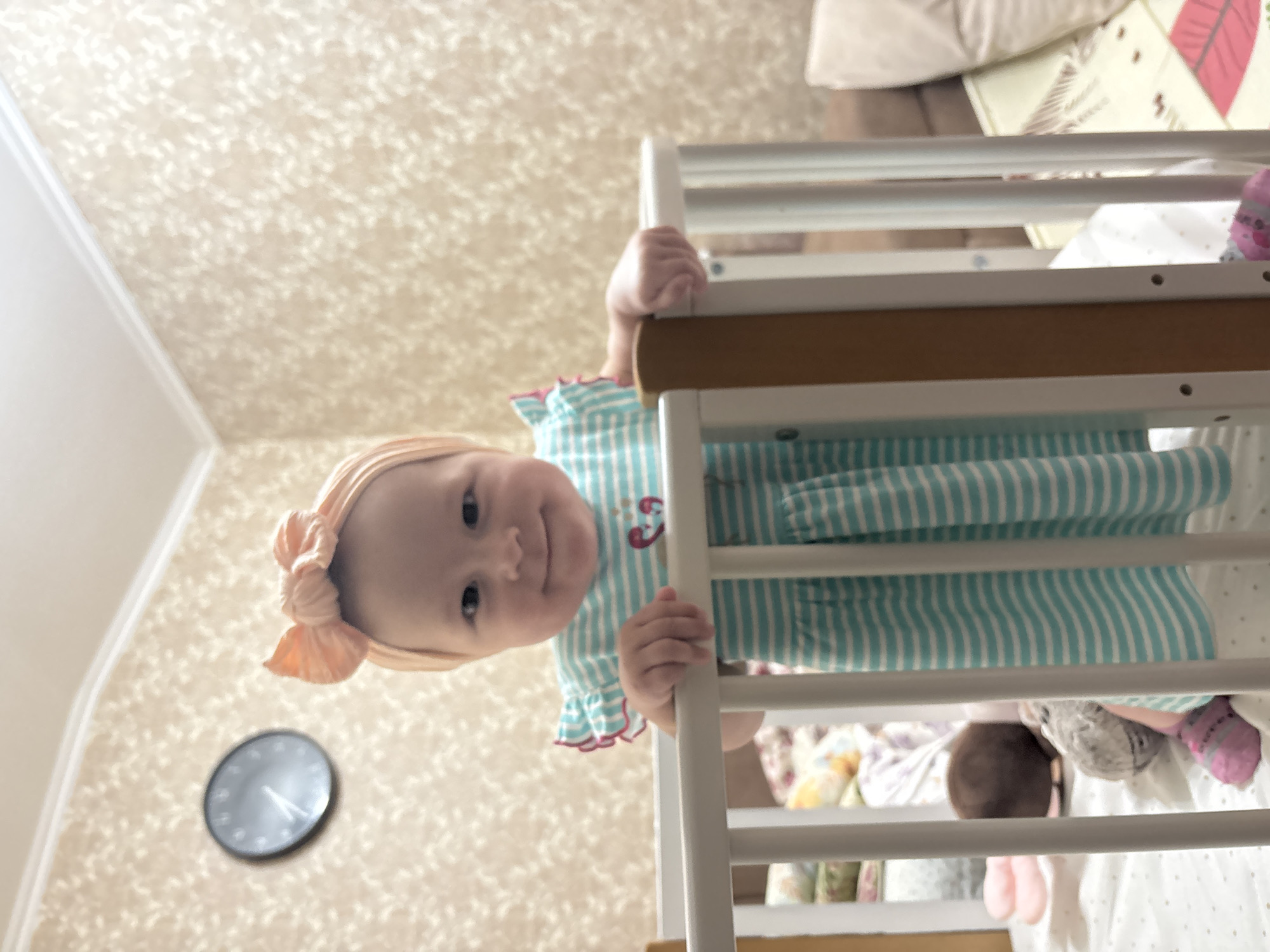 A baby wearing a light peach headband and a turquoise and white striped dress stands in a white crib. The baby is holding onto the side of the crib with both hands and looking at the camera. The baby is wearing pink and purple socks and is looking directly at the camera. A clock hangs on the wall to the left of the crib. The location is Sochi, Russia.