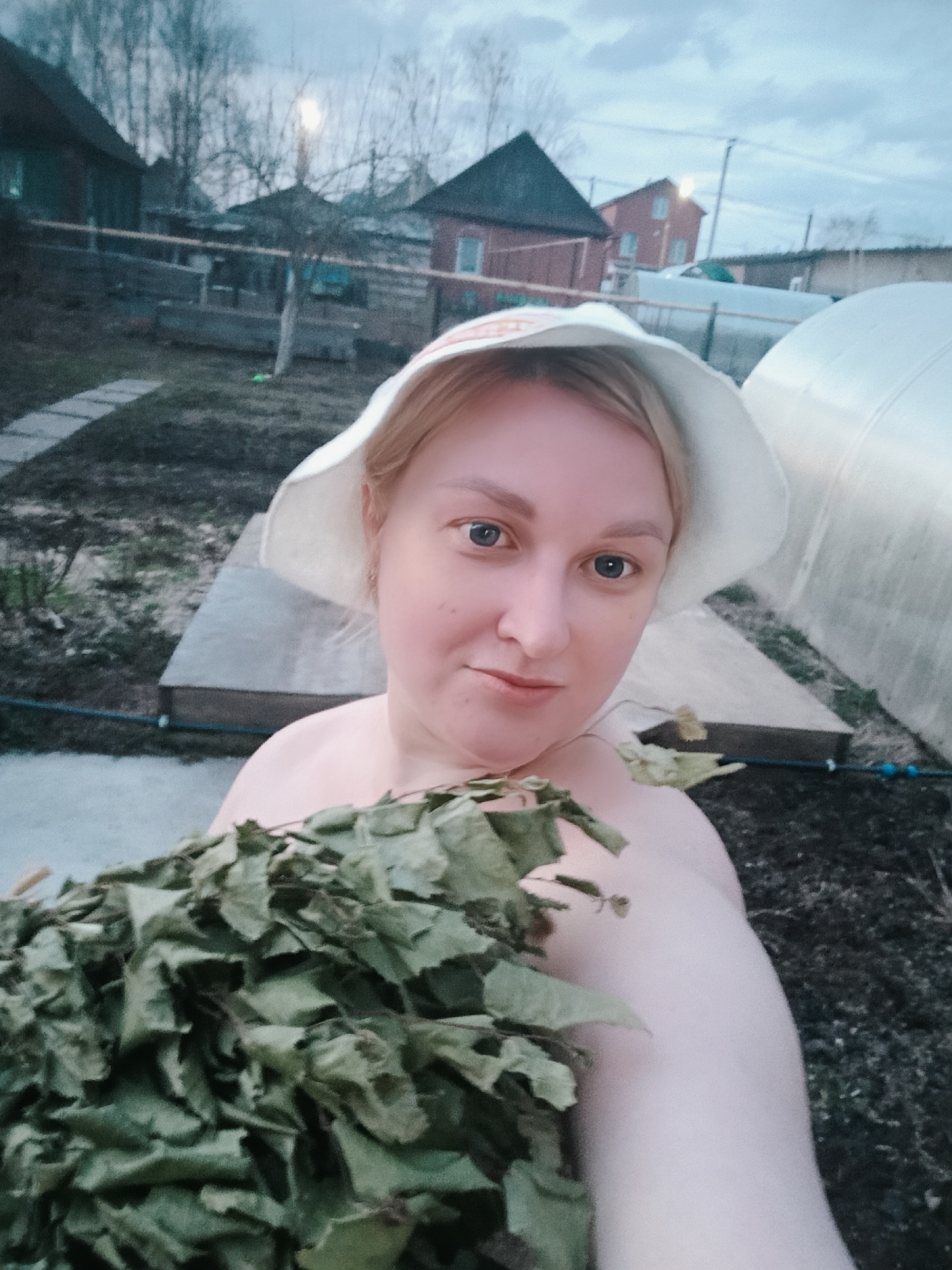 A woman takes a selfie outdoors in a rural or semi-rural backyard in Chaikovsky, Russia. She has fair skin, light hair, and blue eyes, wearing a white hat and appearing topless while holding a large bundle of green, leafy branches, commonly known as a *venik* for a banya (Russian sauna). She looks directly at the camera with a neutral to slightly content expression.
The setting is a garden or yard with various elements. A stone path is visible on the left, leading toward the background. In the mid-ground and background, several houses are present, including a dark-roofed wooden house and a red brick house. Bare trees suggest a colder season like late autumn, winter, or early spring. The sky is overcast, implying a cloudy day, with diffused light that could be late afternoon or early evening.
Other visible objects include a low fence or barrier, dark soil or ground cover, and a large white arched structure resembling a greenhouse on the right. There are also large white tanks or barrels and some dark hoses or pipes on the ground. The scene evokes a rustic, domestic atmosphere, likely related to a banya ritual. No visible text is present in the image.