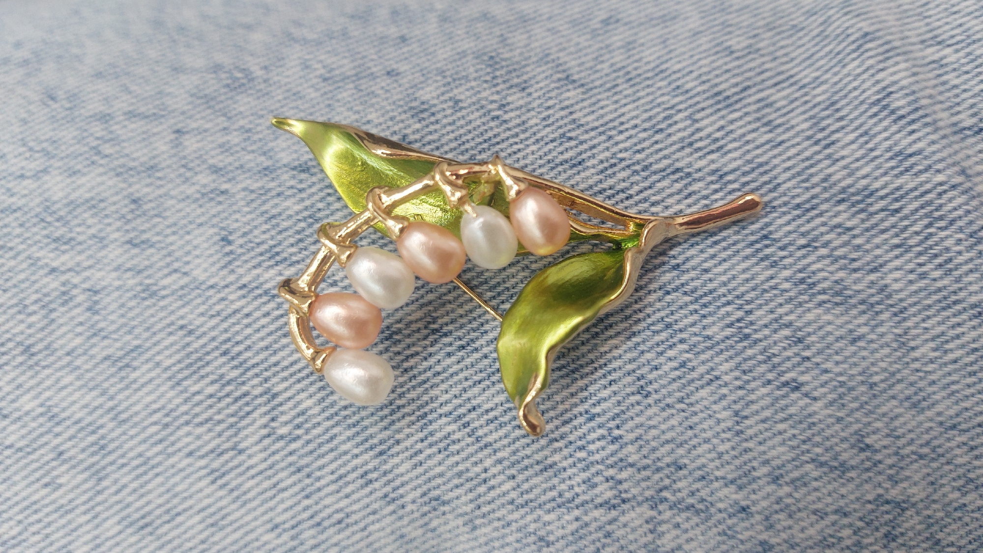 The image depicts a lily-of-the-valley brooch against a blue denim background. The brooch is gold-toned with two green leaves and several small, oval-shaped faux pearls in white and peach hues hanging from the stem. A pin is visible extending from the back of the brooch. The setting is not recognizable as Novosibirsk or any specific city, and there are no people or actions visible in the frame.