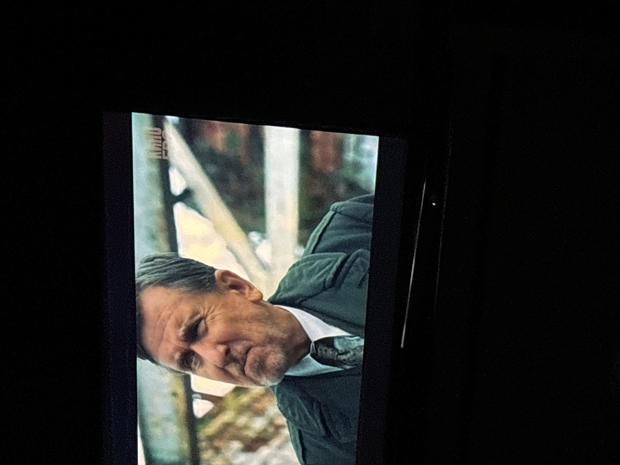 A still image from a television screen shows an older, gray-haired man in a white shirt, dark jacket, and patterned tie in front of what appears to be a dilapidated structure in a snowy or overgrown outdoor environment. The television screen occupies a portion of the left side of the image, surrounded by a black, darkened space. There is a logo or branding along the upper edge of the screen. The man's expression appears stern. The location in Budva, Montenegro is not evident in the image.