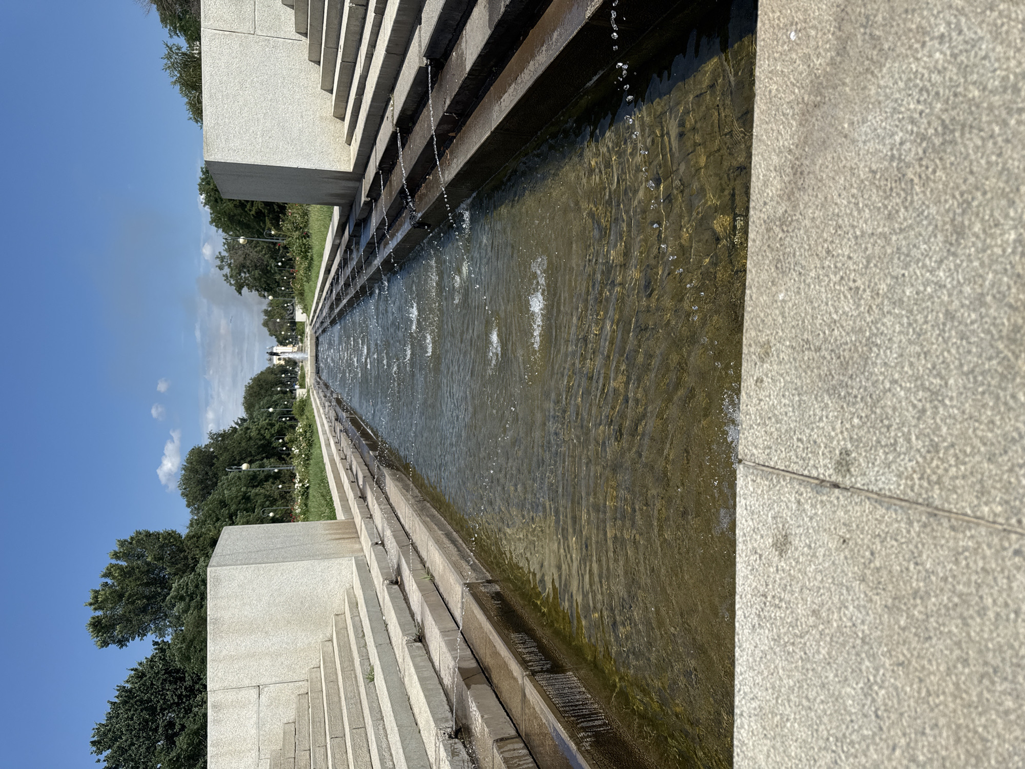 The image depicts a long, narrow water channel with a fountain at its far end, flanked by tiered stone stairs on both sides. Water flows down the channel in cascades, with smaller jets of water spraying from the sides. Trees and greenery line the path beyond the water channel, and a bright blue sky with scattered clouds is visible overhead. The overall setting appears to be an urban park or monument. The location is not Astrakhan, Russia.