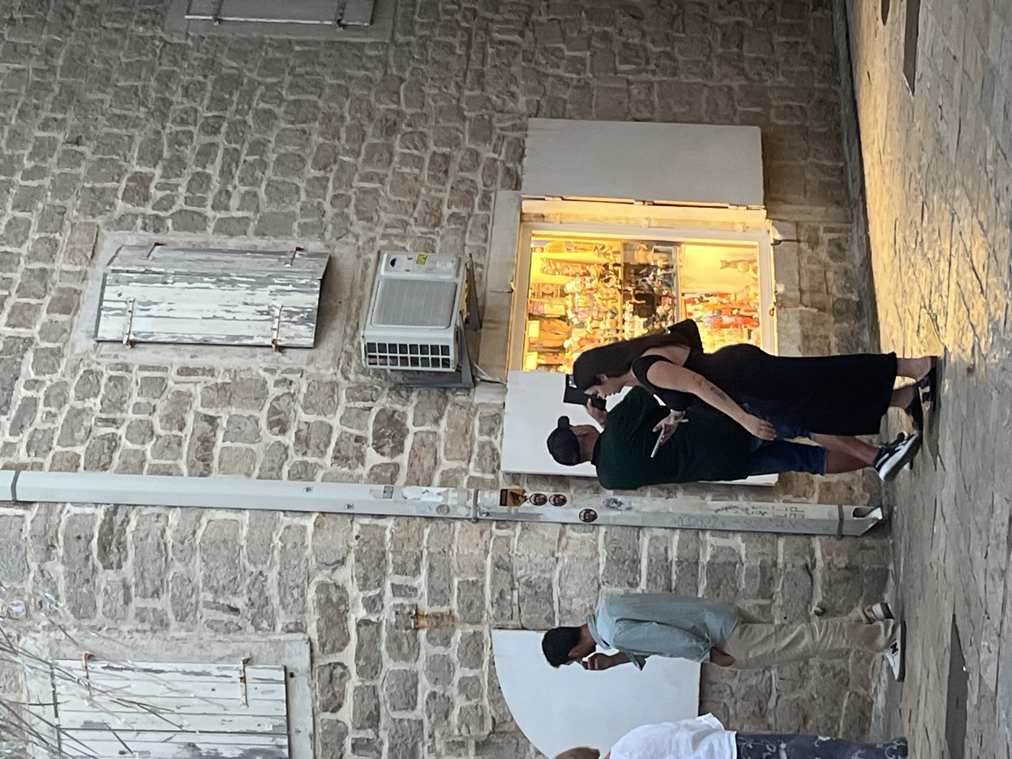 The image depicts a street scene in Budva, Montenegro. A stone building with weathered wooden shutters is visible on the right. In front of the building, a brightly lit shop window displays various items. Three people are interacting in front of the shop. A man wearing a light blue shirt and tan pants stands on the left, holding a phone to his ear. To his right, a man in a dark green shirt and blue shorts, wearing a baseball cap, is holding a phone and appears to be taking a picture. A woman in a black dress stands next to him, also looking towards the shop. The ground is paved with cobblestones, and the lighting suggests it is either early morning or late afternoon.