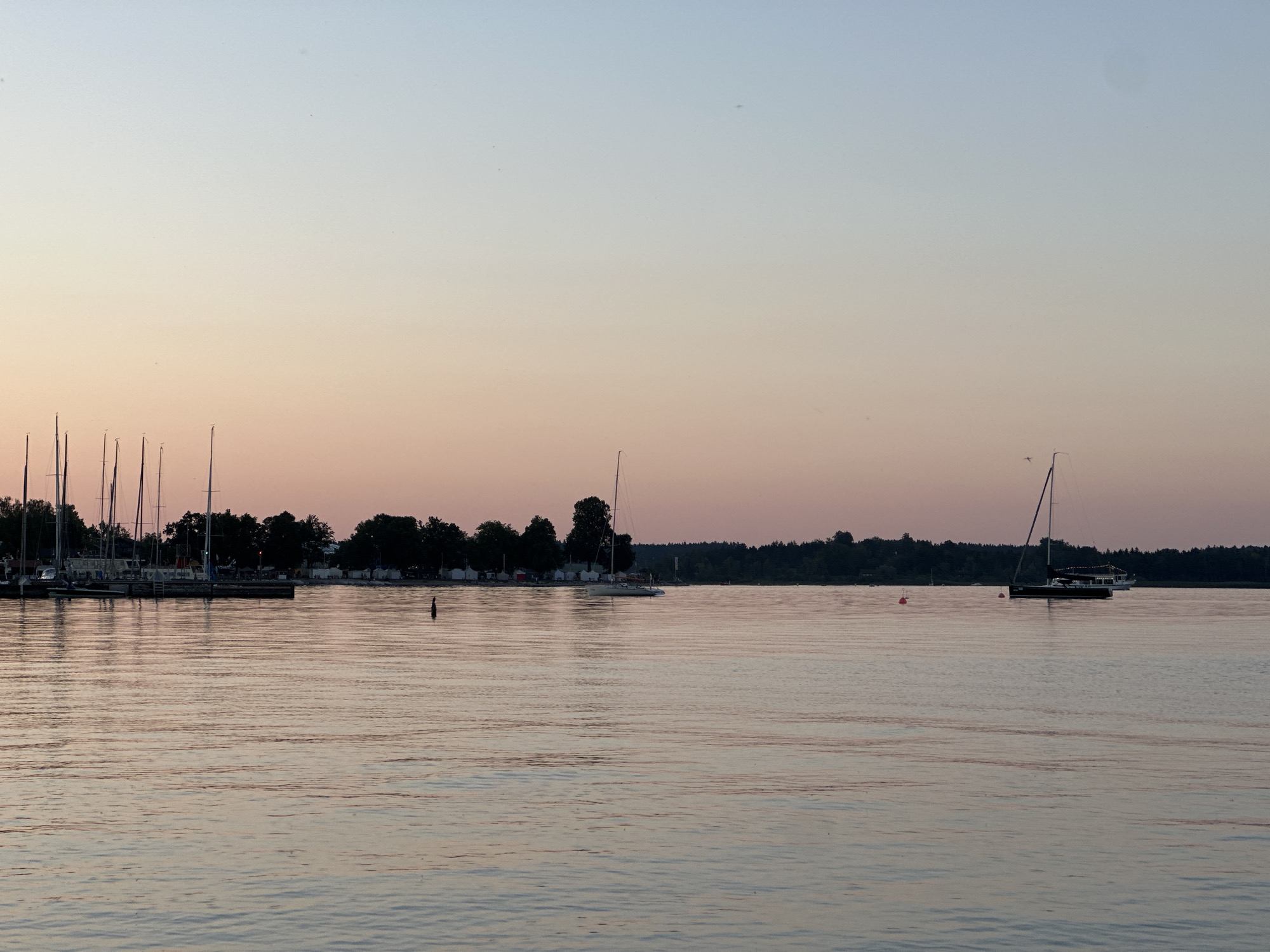 The image depicts a tranquil scene at a marina in Prien am Chiemsee, Germany, during twilight. The sky transitions from a pale blue at the top to a soft peach and pink hue near the horizon, indicating the sunset or sunrise. The calm water of the lake reflects the subtle colors of the sky and the silhouettes of various boats.
In the foreground, the rippling water is the dominant element, with gentle waves creating a textured surface that catches the fading light.
In the mid-ground, a marina is visible along the left side of the image, featuring a row of sailboats with their masts standing tall against the sky. Some of these sailboats appear to be docked. Further out on the water, a single white sailboat with a tall mast is sailing to the right, its hull dark against the water. To the right of this sailboat, a larger, dark-hulled boat, possibly a motor yacht, is also visible. A red buoy is present in the water to the left of the dark-hulled boat.
In the background, a treeline of dark green foliage forms a dense border along the far shore. Behind the marina on the left, a few white tents or awnings suggest a campground or event area. A single red buoy is positioned in the water in front of the treeline and to the left of the marina. The overall atmosphere is serene and peaceful, characteristic of a quiet evening by the lake. There are no visible people in the frame.
