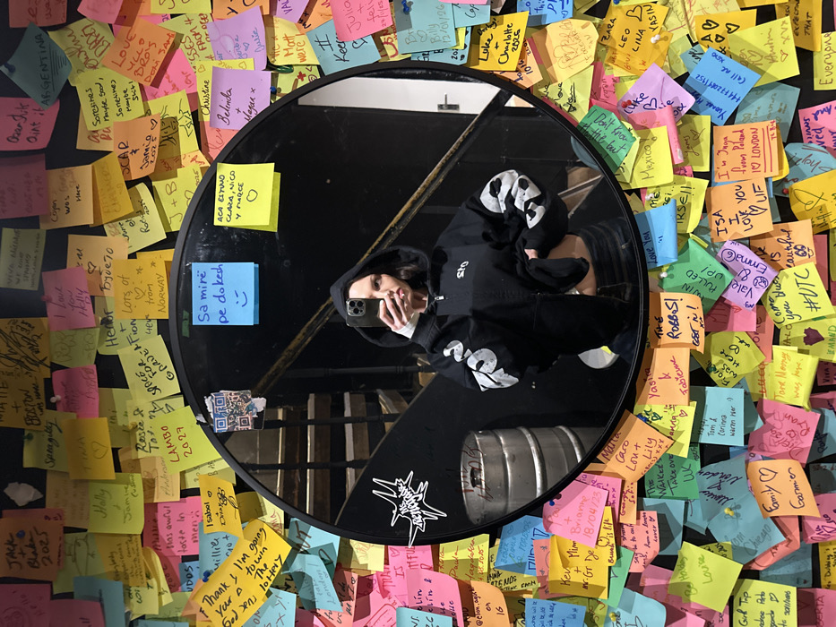 A young adult female is depicted in a mirror selfie taken within a location likely to be The Old Blue Last in Shoreditch, London, United Kingdom. The subject, visible from the mid-thigh up, wears a black hooded zipper jacket with white graphic designs on the sleeves and "STO" on the left chest; the jacket is partially unzipped, exposing her midriff. She holds a silver-colored smartphone with both hands, angled to capture her reflection, which partially obscures her face. The large, circular mirror, framed in black, is surrounded by a wall completely covered in hundreds of multicolored Post-it notes, each containing handwritten messages, names, dates, or drawings. Specific notes on the mirror itself include "Sa mirë pe dokesh" (Albanian for "How beautiful you look") with a smiley face, and another yellow note with names and a heart. A white star-shaped sticker and a QR code are affixed to the mirror. The mirror's reflection reveals a dark interior background, including a visible staircase with dark treads and a handrail, and a silver metal keg positioned at the base of the stairs.