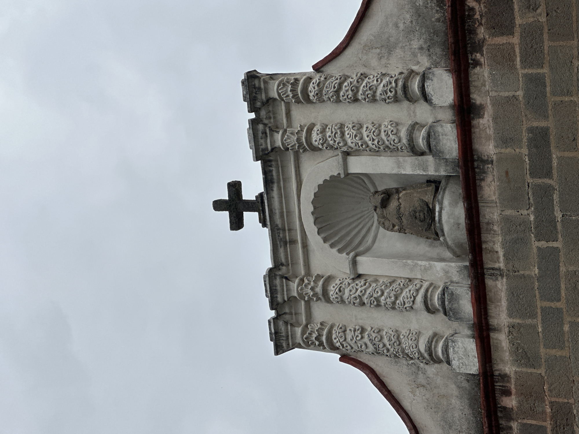 A close-up, low-angle shot depicts the upper section of a building, potentially a church, located in San Miguel Zinacantepec, Mexico. The architecture is characterized by grey stone and ornate plasterwork. A simple black cross is prominently positioned at the top center. Below the cross, a niche is visible, containing a weathered stone bust or statue. The building's facade features decorative columns with intricate floral or vine-like carvings. A brick wall abuts the architectural structure on the right side of the frame. The overcast sky provides a neutral backdrop.