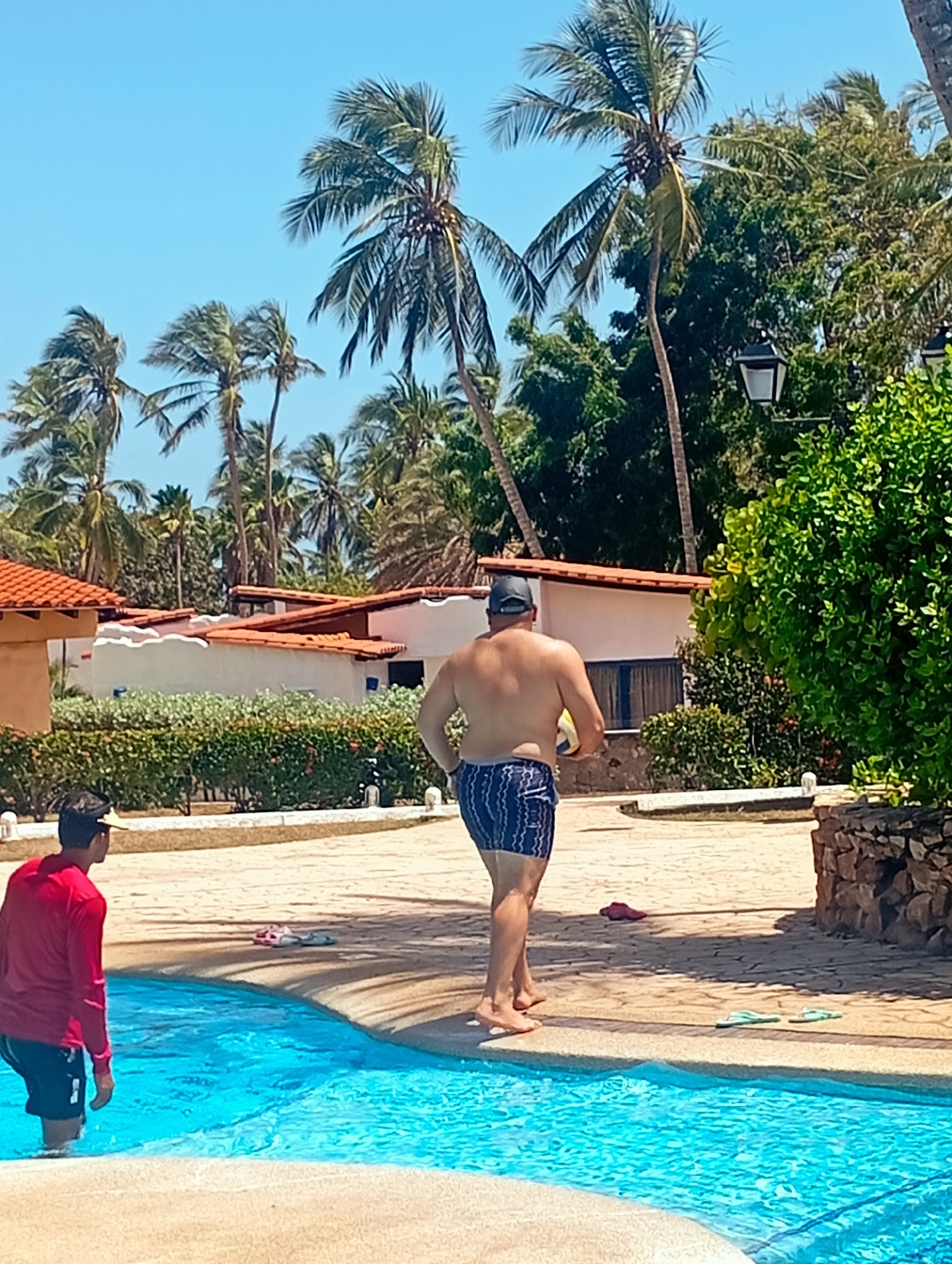 The image captures a scene at a resort in Playa El Agua, Venezuela. The scene is sunlit with a bright blue sky filled with palm trees. A man wearing blue swim trunks and a dark baseball cap is walking on a tiled patio away from the pool, holding a yellow volleyball. Another person, wearing a red long-sleeved shirt and dark shorts, is standing in the pool on the edge, facing away from the camera. Scattered on the patio, near the pool's edge, are pairs of flip-flops. In the background are white buildings with orange tile roofs, separated by green hedges. A dark green bush is on the right.