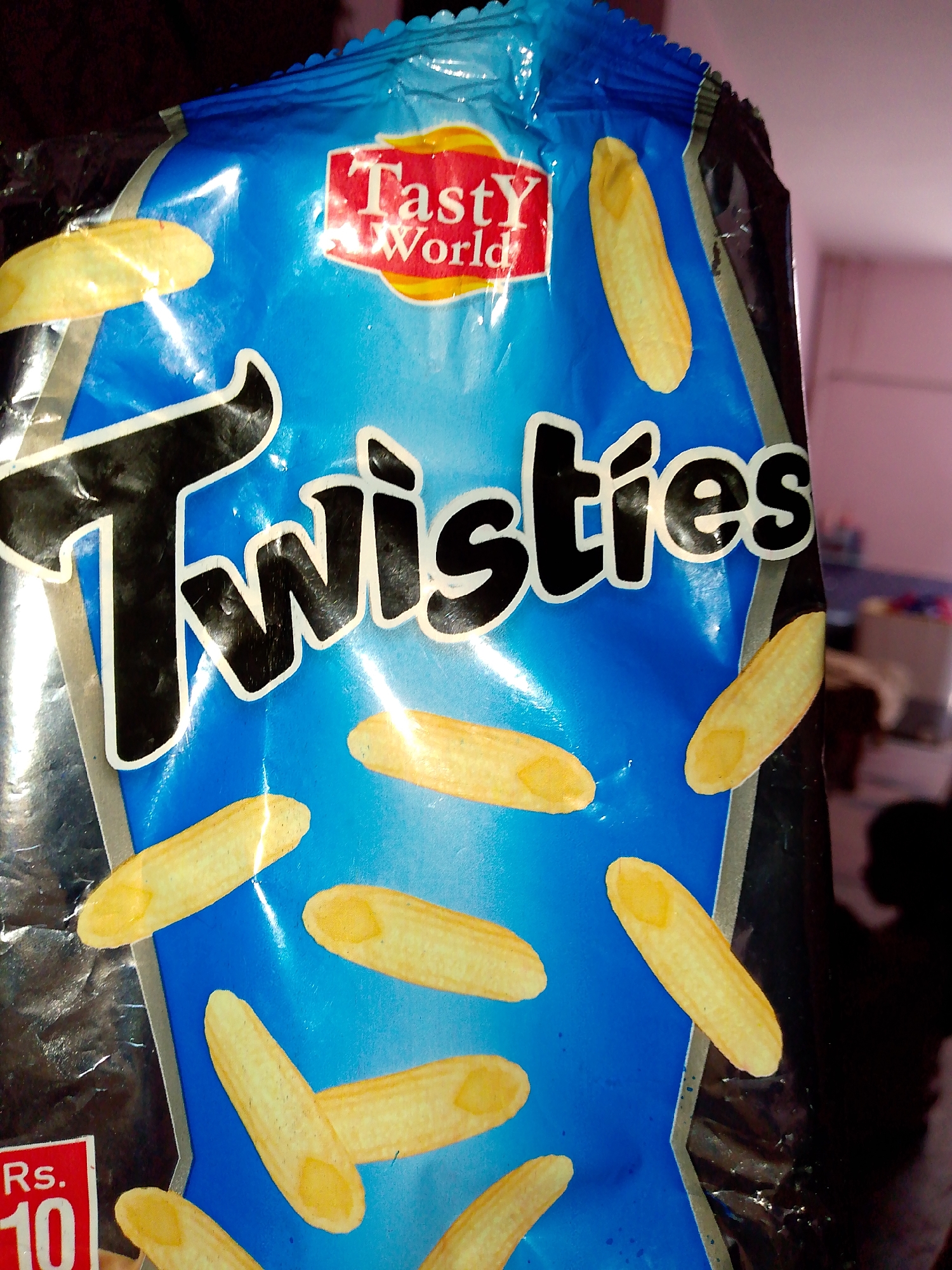 The image shows a close-up view of a blue snack bag with the brand name "Twisties" prominently displayed in large, black, stylized letters with a white outline. Above "Twisties" is a red logo that says "Tasty World". Several elongated, yellow, textured snack pieces are scattered across the blue background of the bag. In the bottom left corner, a red price tag displays "Rs. 10". The bag appears to be made of a crinkled, shiny plastic material. The background is dark and out of focus, suggesting the photo was taken indoors with limited lighting. The overall scene is a product shot of a snack food item. There is no information about the city or location.
