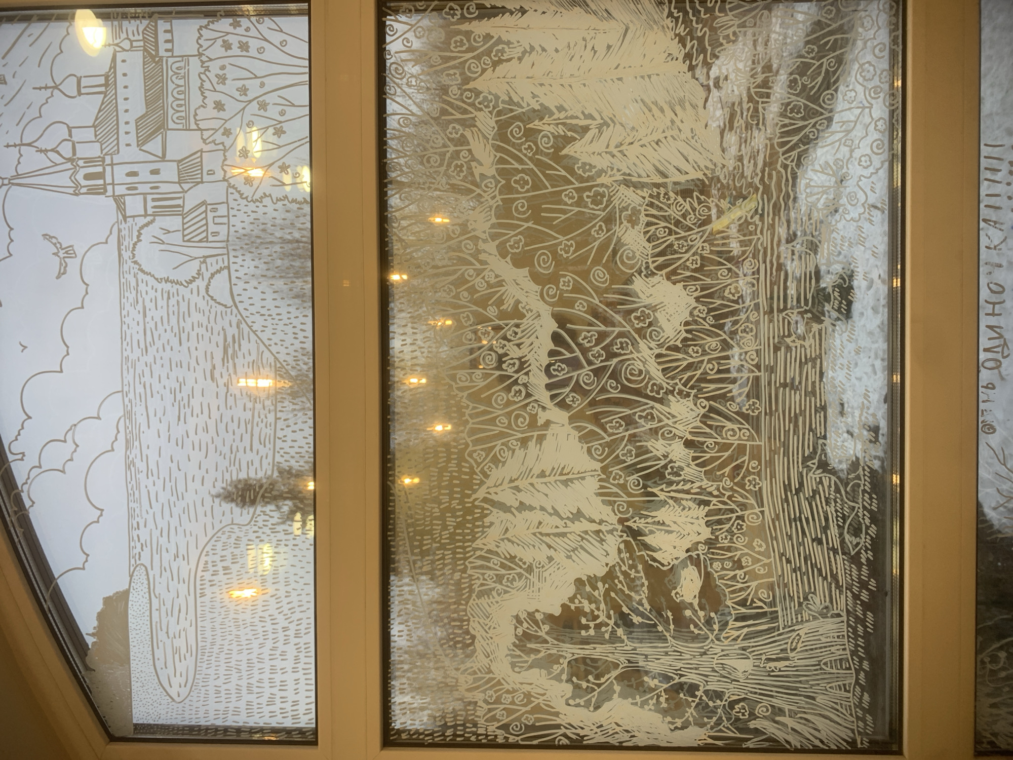 The image shows two adjacent window panels with etched drawings, part of a series of windows. The left panel features a sketch of Russian Orthodox architecture, including a church with multiple domes and a bell tower, set in a landscape with a bird. The right panel displays a detailed depiction of a forest scene, including trees, foliage, and possibly figures near the bottom. The windows are framed in a light brown material, likely wood or a wood-like composite. The building's location is in Izhevsk, Russia. The etchings on the glass create a contrast between the light etched areas and the transparency of the glass, which offers a glimpse of the scene outside.