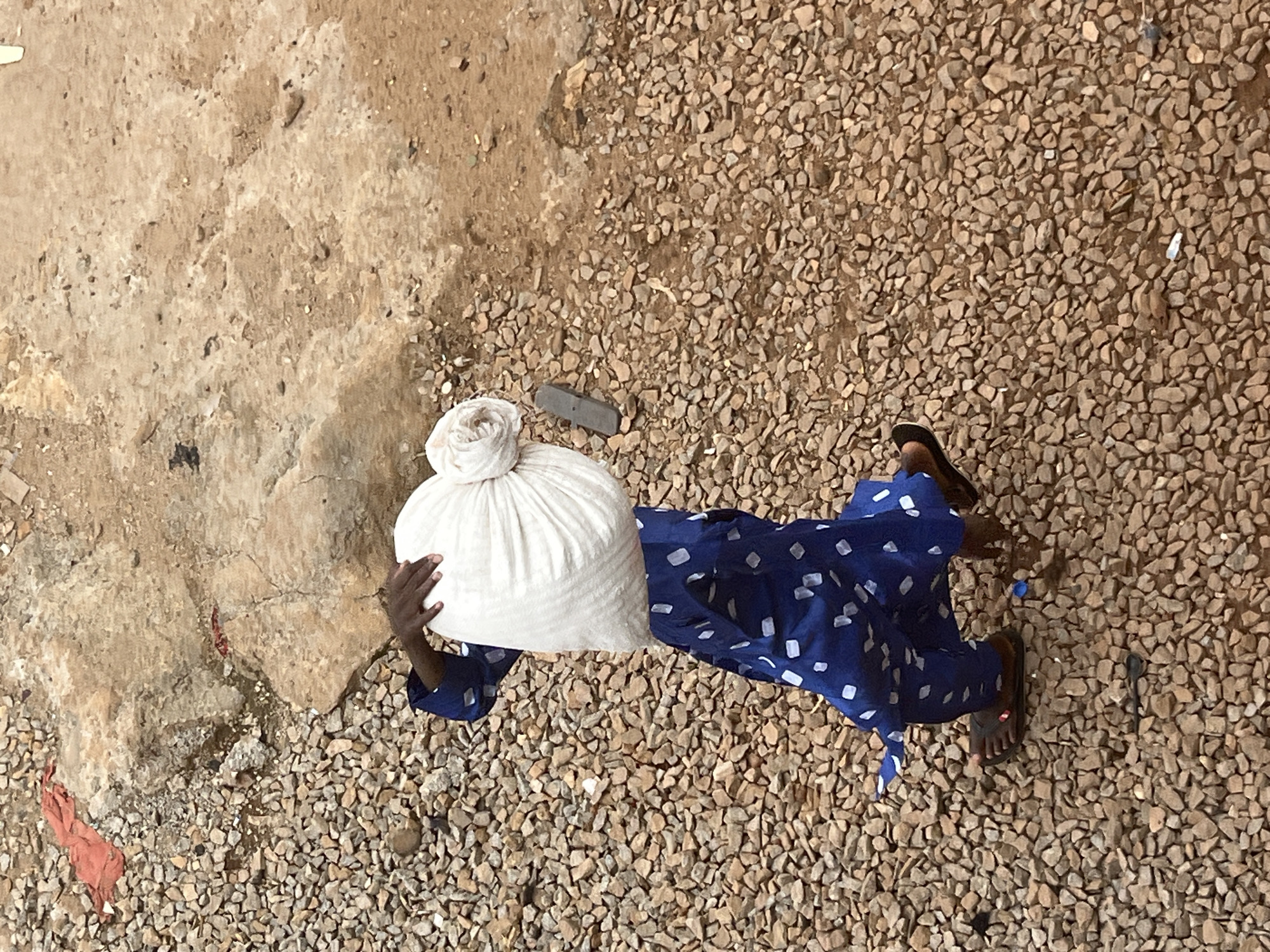 An aerial view captures a person walking on a gravel-covered ground in Jos, Nigeria, carrying a large, white bundle on their head. The individual is wearing a blue garment adorned with white polka dots and brown sandals. The surroundings consist of loose rocks and some dirt.