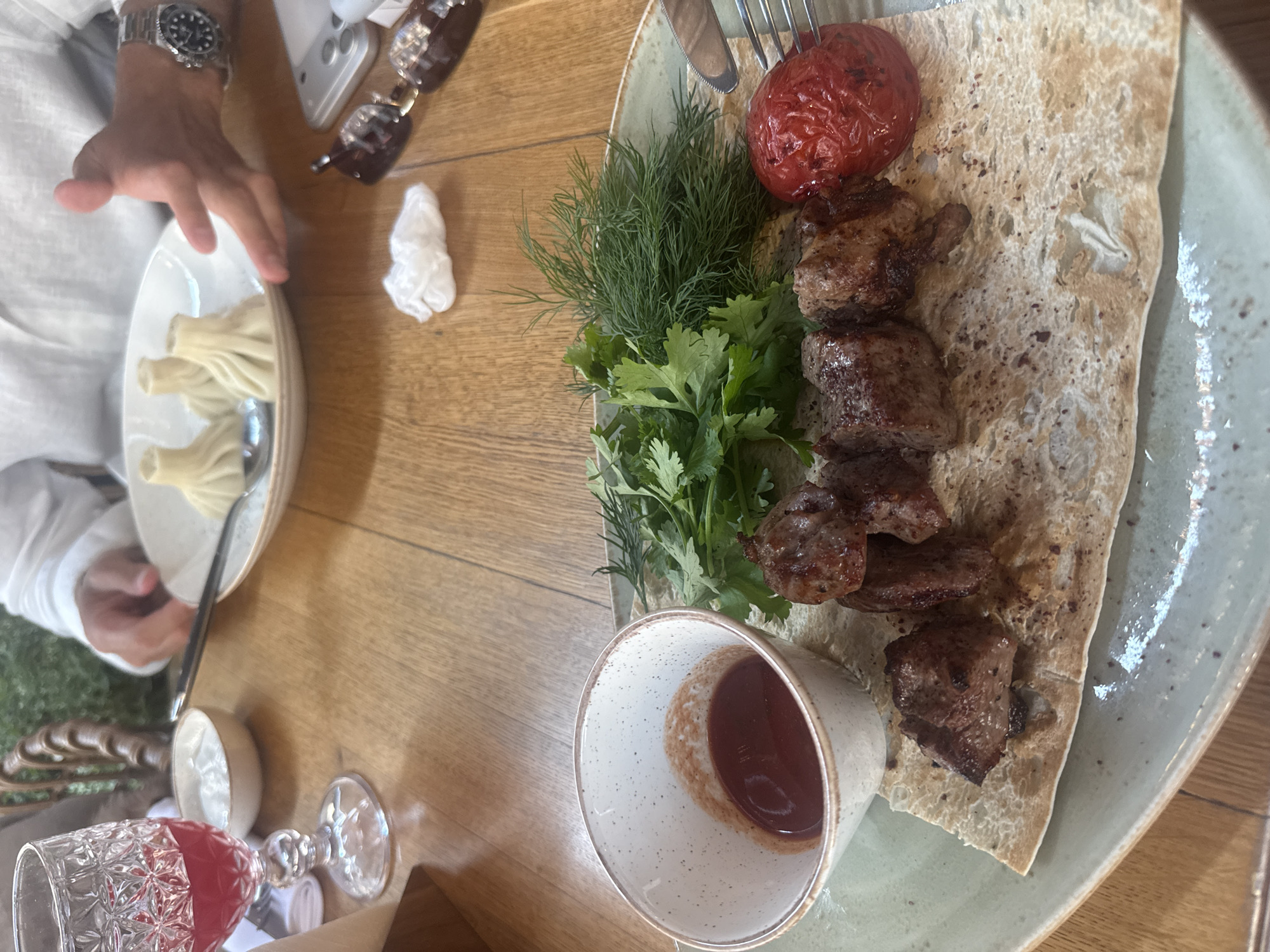 A close-up, eye-level shot shows a meal on a wooden table, likely in a restaurant in Moscow, Russia. In the foreground, a light green plate holds a serving of grilled meat skewers, a whole roasted tomato, fresh herbs (cilantro and dill), and a small bowl of red sauce. The meat is skewered and sits on a piece of flatbread. In the background, partially visible, are plates of khinkali dumplings, a glass of red liquid, a small bowl of white sauce, and a person's hands, one wearing a wristwatch. Sunglasses and a phone are also on the table. The environment appears to be indoors, with diffused lighting.