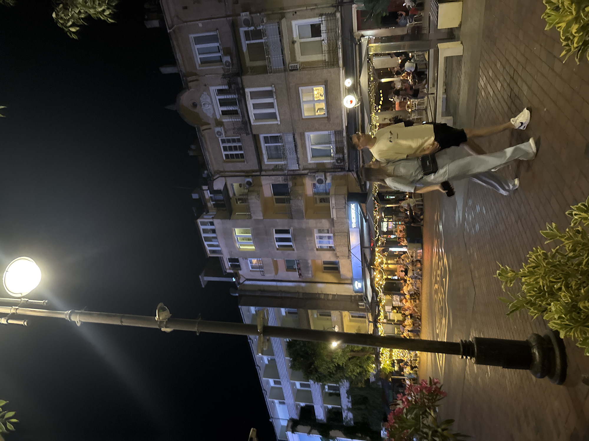 At night on a pedestrian street in Varna, Bulgaria, a couple walks hand-in-hand past a row of illuminated restaurants. The man wears a light-colored t-shirt and black shorts, while the woman is dressed in a light blue coordinated set. They are walking on a brick sidewalk. Behind them, buildings with multiple stories are visible, featuring windows, balconies, and air conditioning units. Several restaurants have outdoor seating areas decorated with string lights, and patrons are seated at tables. A streetlamp with a bright light is positioned in the foreground, and foliage can be seen at the bottom of the frame. The sky is dark.