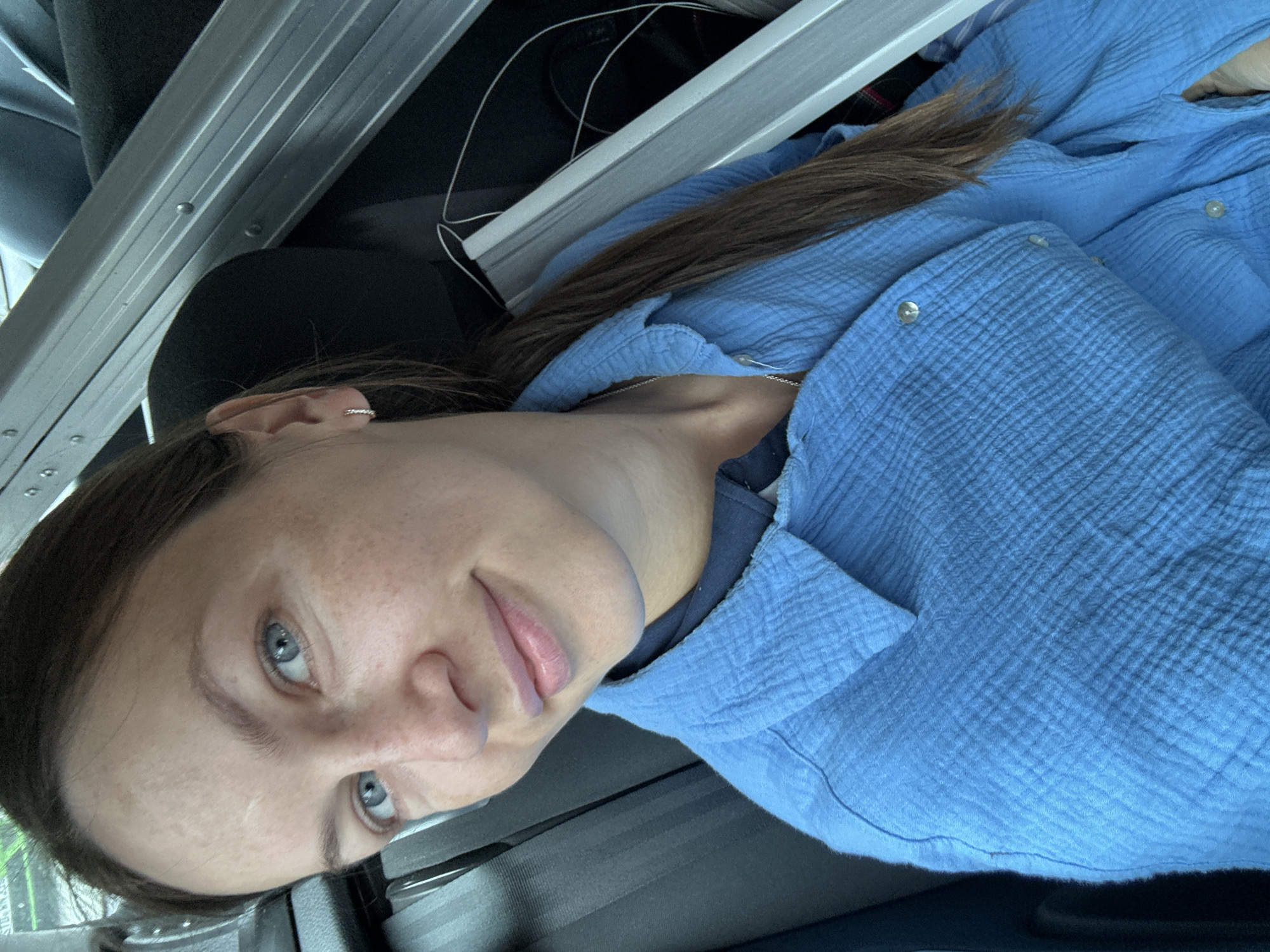 A woman with light skin, blue eyes, and brown hair pulled back, with a section draped over her left shoulder, is depicted in a medium close-up, high-angle portrait within a vehicle in Deinste, Germany. Her face shows visible freckles and a faint linear mark on her forehead. She is looking directly at the camera with a slight smile, revealing light pink lips. A small, silver-colored hoop earring is visible in her right ear. She wears a textured, light blue, long-sleeved button-up shirt over a darker blue collared garment, and a thin silver chain necklace is visible around her neck. A dark grey seatbelt crosses her left shoulder. In the background, to her right, multiple long, silver-colored metallic objects with visible rivets are positioned on a dark surface, potentially a rear seat, alongside a white cable. The visible interior of the vehicle is dark grey and black.