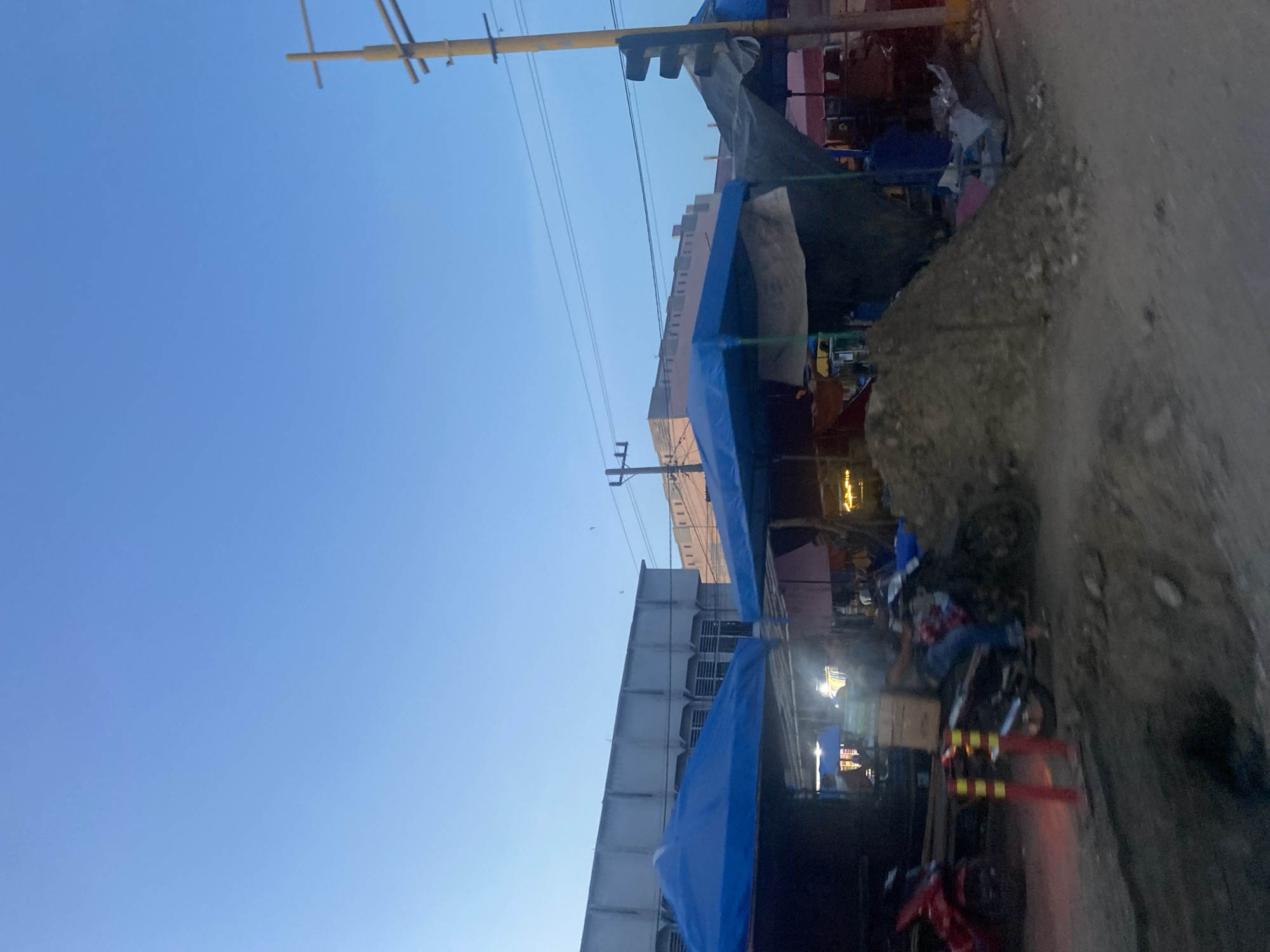 The image depicts a street scene in Keudelapang, Indonesia. The view is angled upward, showcasing a clear blue sky above buildings and street-level activity. Power lines stretch across the sky. Buildings are visible, some with blue tarps or awnings extending outward. A dirt or gravel embankment runs along the right side of the frame. Motorcycles are parked and possibly in use on the street. Red and yellow barricades are present. A pile of dirt or other material sits beside the street. The scene appears to be in a commercial or residential area with a mix of formal buildings and temporary structures.