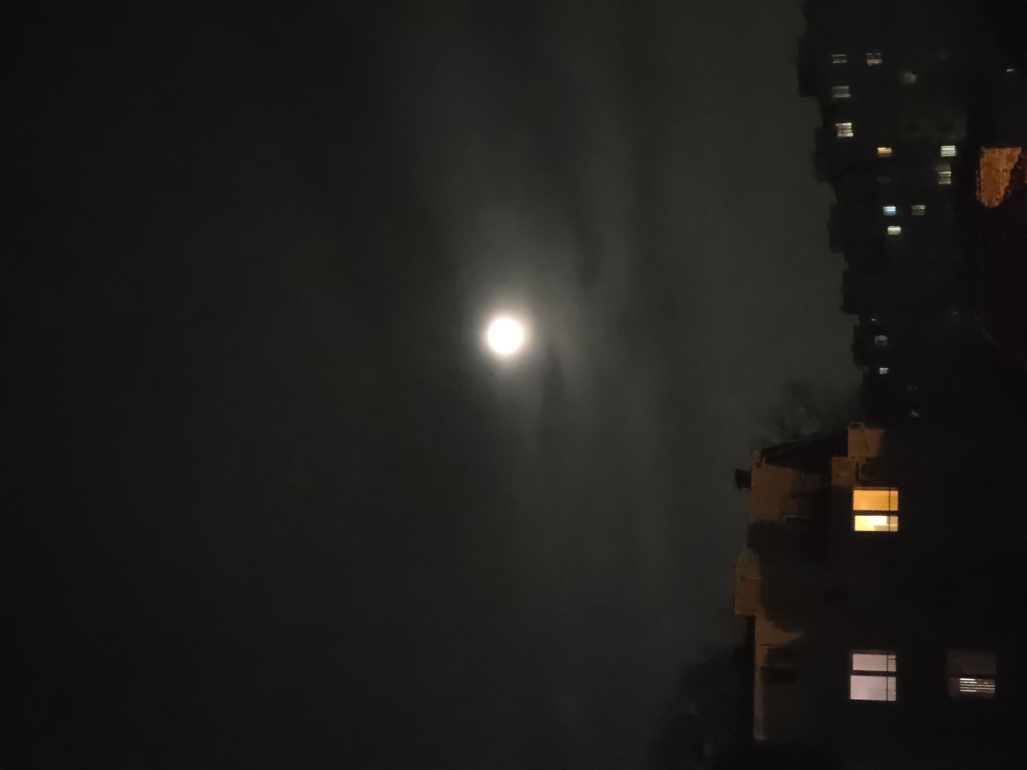 The image shows a night scene with a bright, full moon partially obscured by clouds in the sky. The moon is located towards the left side of the frame. On the right, there are dark buildings, likely residential apartments, with a few illuminated windows suggesting occupancy. The overall environment suggests an urban setting at night. The photograph's composition focuses on the contrast between the moon's luminescence and the darkened buildings. The specific location in Varna, Bulgaria cannot be determined based on the image alone. There are no people visible in the image.