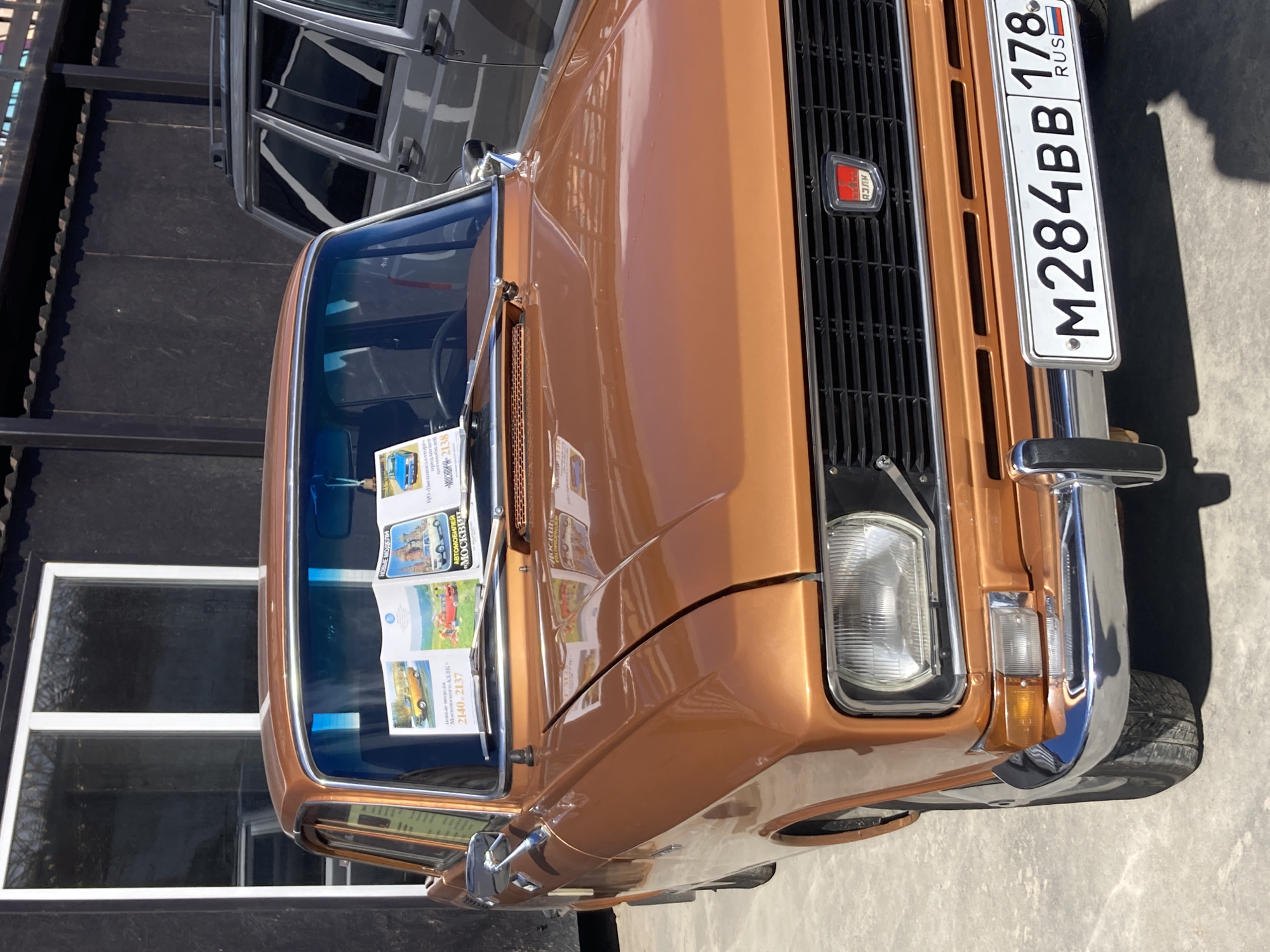 The image shows the front end of a brown-colored vehicle, specifically a vintage car, with its grill prominently displayed. The license plate reads "M284BB 178," indicating it's a Russian vehicle. A car model emblem "АЗЛК" is on the grille. A set of books are placed on the dashboard. The picture shows a car from the city of Saint Petersburg, Russia.