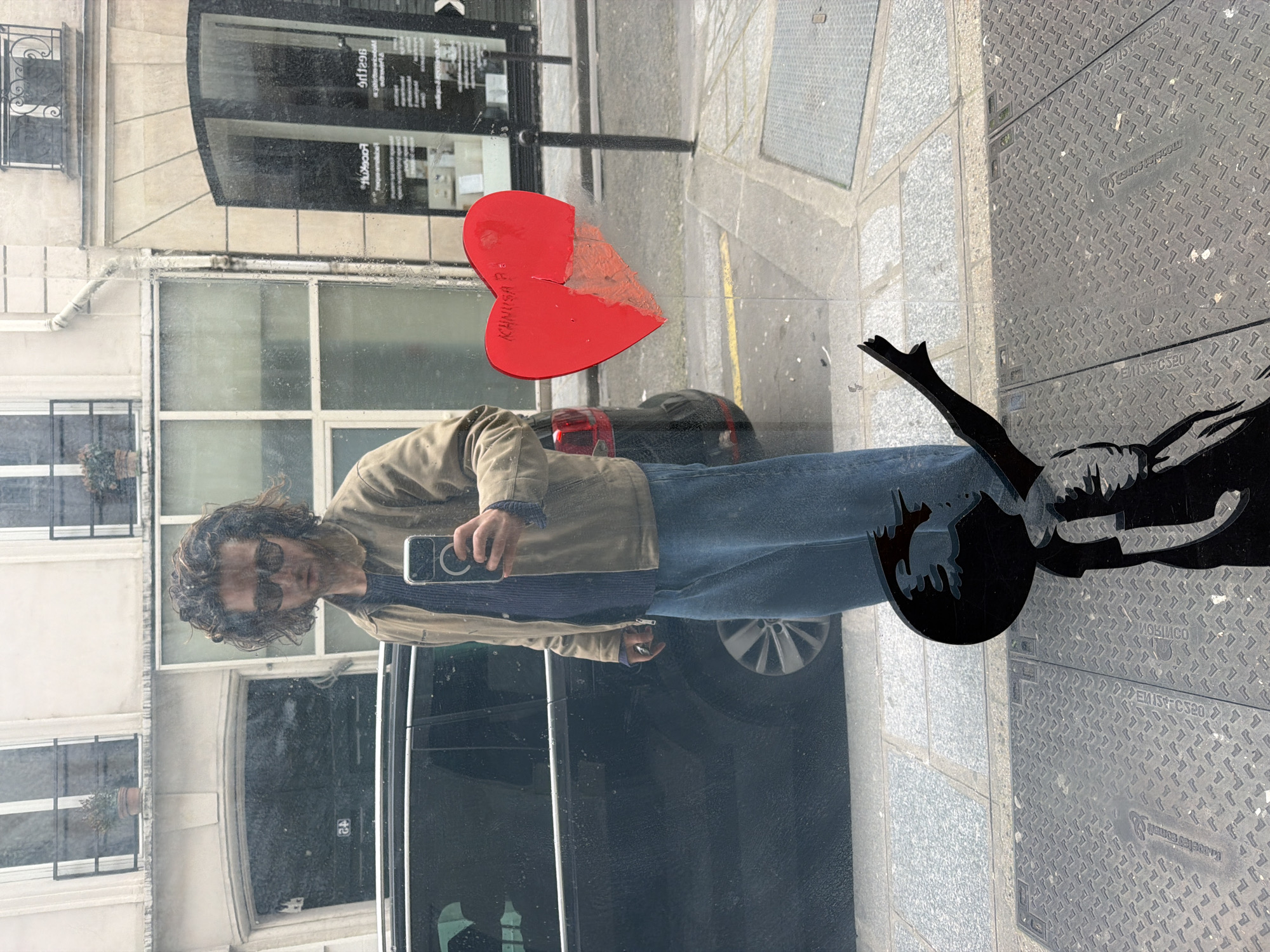 The image shows a reflection in a window, likely a shop window, of a person taking a picture with a phone, the street, and the building across the street. The person has brown hair and is wearing sunglasses, a blue shirt and a beige jacket. The reflection shows a piece of red paper in the shape of a broken heart stuck to the window. The word "MANUSA" is written on the heart. Outside the window, a black metal street decoration, and the sidewalk can be seen. The building reflected in the window is light-colored with multiple windows. This location is within the city of Paris, France.