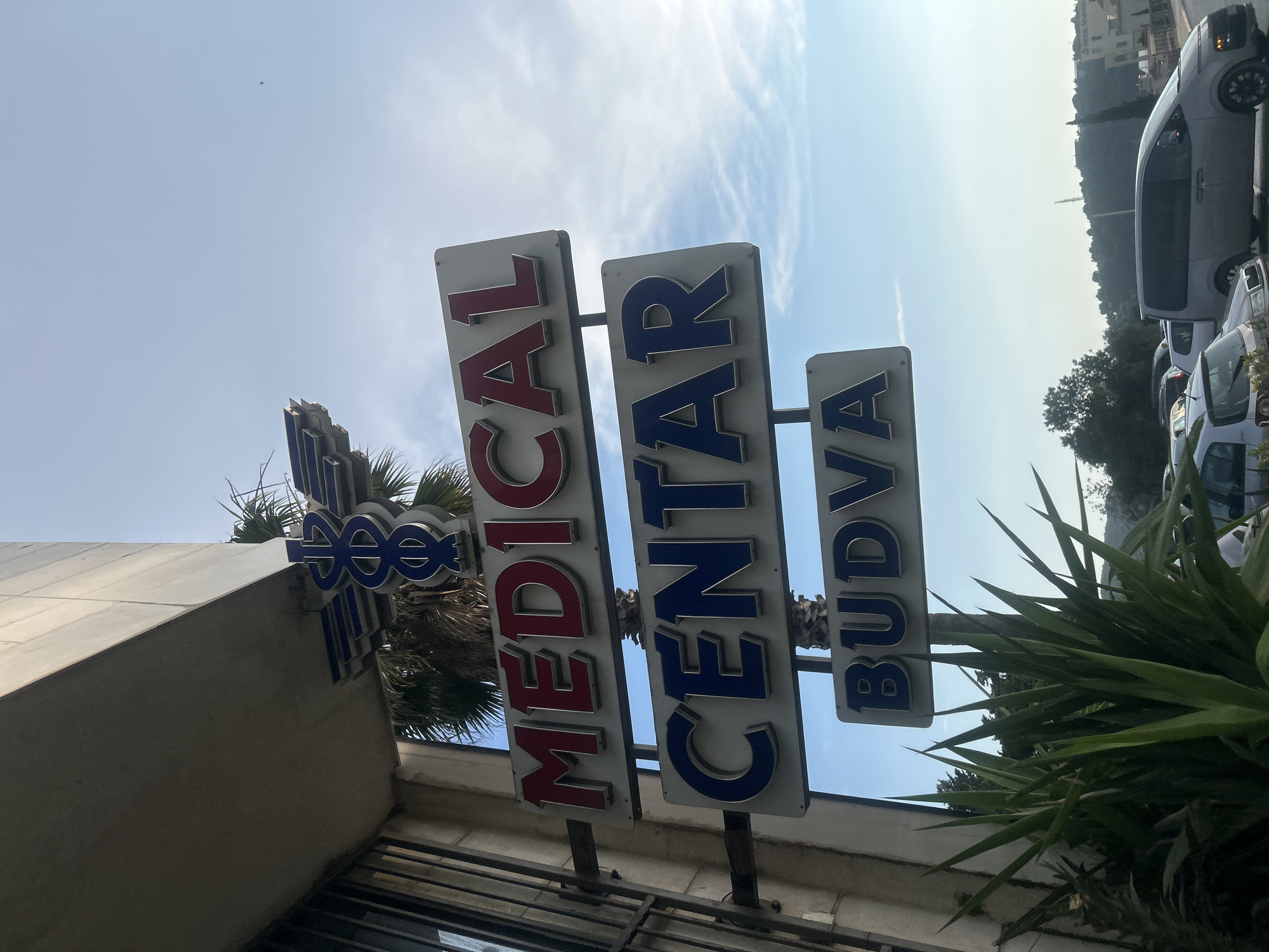 The image shows a sign for "MEDICAL CENTAR BUDVA" in Montenegro. The sign has three vertical panels with raised lettering. The first panel reads "MEDICAL" in red letters with a blue outline. The second panel reads "CENTAR" in blue letters with a red outline. The third panel reads "BUDVA" in blue letters with a red outline. Below the "MEDICAL" panel is a blue logo that appears to be a stylized caduceus, a symbol of medicine.
The sign is mounted on poles in front of a building with a light-colored facade and some windows. Palm trees are visible behind the sign. In the background, there are some vehicles, including a white van and other cars parked in what appears to be a lot. Further in the background, a hillside is covered with trees and buildings, and a tall, slender structure, possibly a minaret, is visible on the hilltop.
The sky is bright blue with some wispy white clouds, suggesting it is daytime and the weather is clear. The sun appears to be high in the sky, casting some light and shadows. The overall impression is that of a sunny day in a coastal or urban area. The perspective of the photo is tilted upwards, showing the sign against the sky.