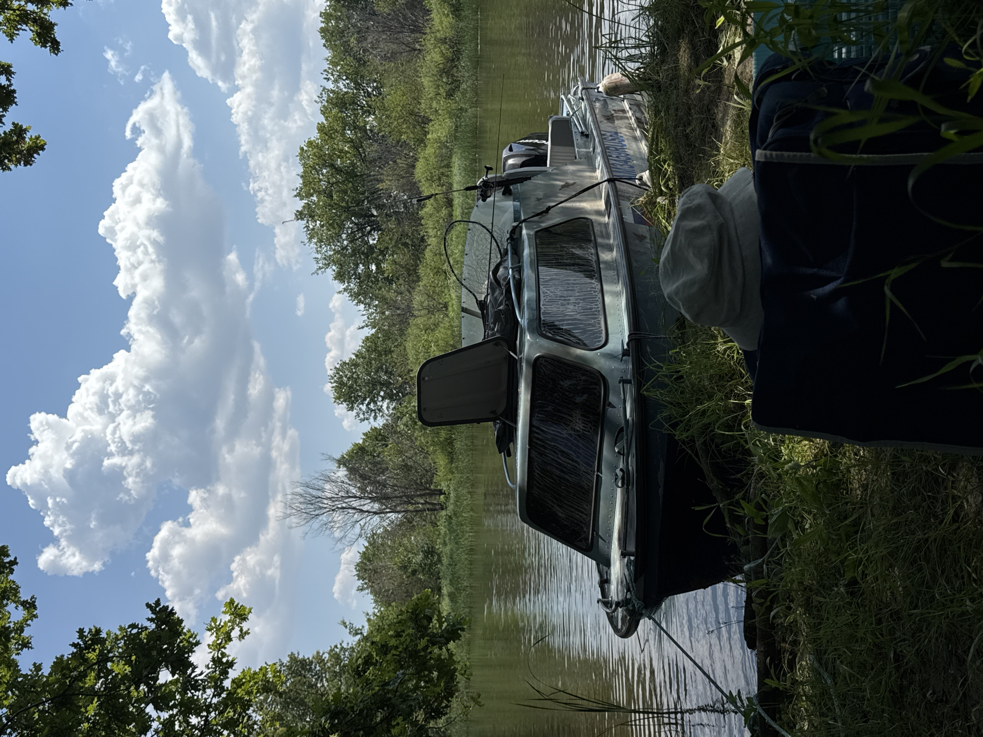 A silver boat is moored on the bank of a wide, calm river. The boat has a cabin with tinted windows, a fishing rod, and a net visible. Fishing equipment is also attached to the boat. On the riverbank, a chair is set up, on which a light-colored hat rests. The bank is covered with green grass and reeds. Trees and greenery line the opposite bank of the river. The sky is blue with fluffy white clouds. The scene is located in Krasnyi Yar, Russia.