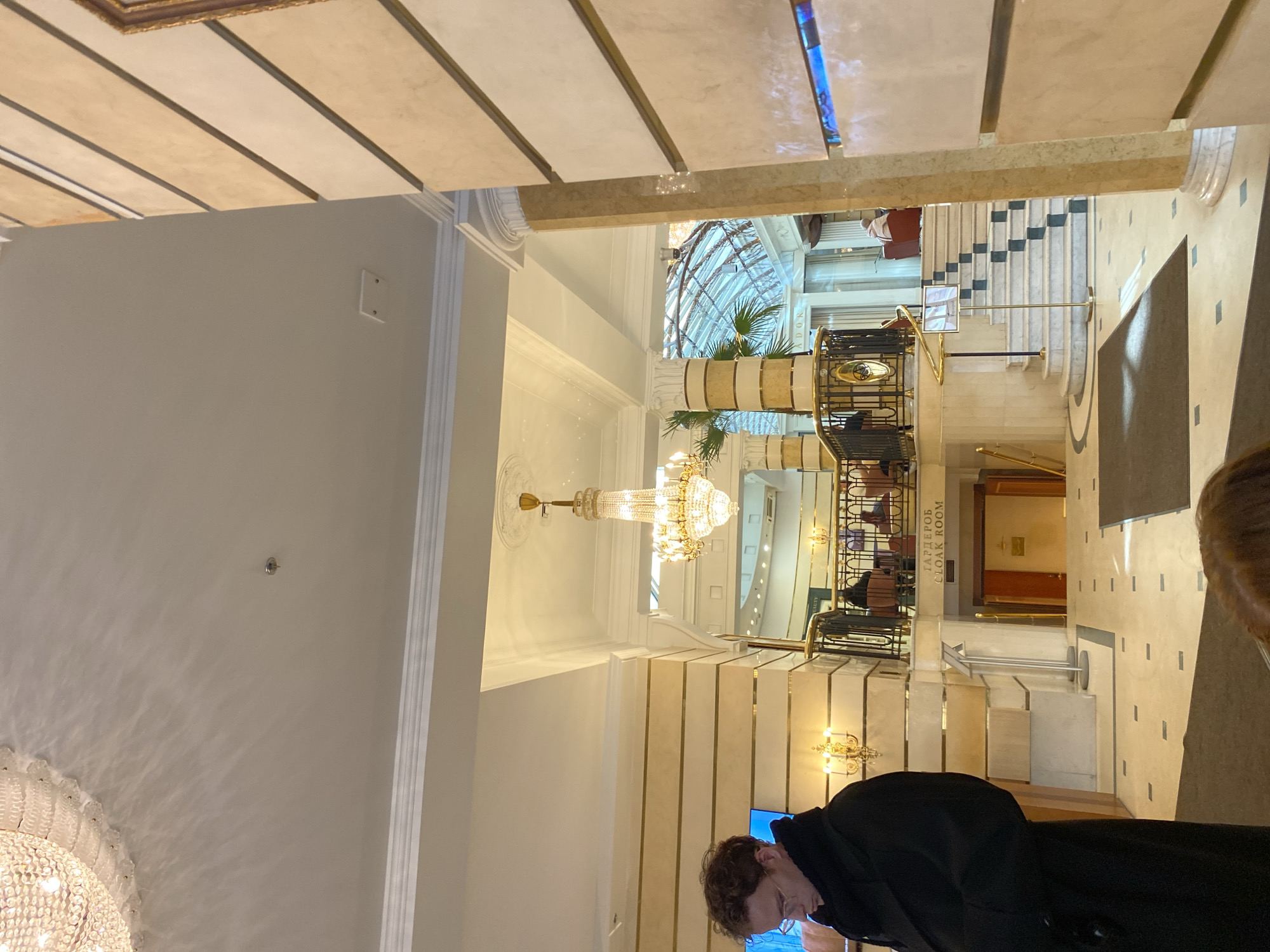 The image depicts a luxurious multi-level interior, likely a hotel lobby or grand public building, located in Saint Petersburg, Russia.
The environment features classical architectural elements, including light-toned walls with horizontal gold-colored banding, white ornate crown molding, and a high ceiling from which multiple large crystal chandeliers are suspended. A fire sprinkler head is visible on the ceiling in the upper left. The flooring consists of cream-colored tiles with darker square inlays, partially covered by dark rectangular rugs. A prominent marble-clad pillar frames the scene on the right, adorned with horizontal light and dark brown striped panels and a narrow vertical blue illuminated strip.
In the foreground, a young adult male with light brown, curly hair and glasses is positioned on the left, wearing a dark overcoat. He is seen in profile, facing slightly downwards and to the right, appearing to look at an object in his hands or on a display. The top of another person's head with reddish-brown hair is visible in the extreme bottom foreground, partially out of frame.
The middle ground reveals a mezzanine level with a black and gold balustrade, where several individuals are seated at tables, largely indistinct. One person in a red armchair is visible on the mezzanine. Below the mezzanine, a doorway marked with "ГАРДЕРОБ CLOAK ROOM" in both Cyrillic and English indicates a cloakroom entrance to a lower level. To the right of this, a grand curved marble staircase with a matching black and gold balustrade descends. An information stand with a display is placed near the stairs. Further in the background, beyond the mezzanine, a large glass-domed roof structure is visible, through which natural light appears to filter. Large decorative columns with potted palm trees are also visible on the mezzanine level. Wall sconces with candle-like bulbs provide additional illumination throughout the space.
