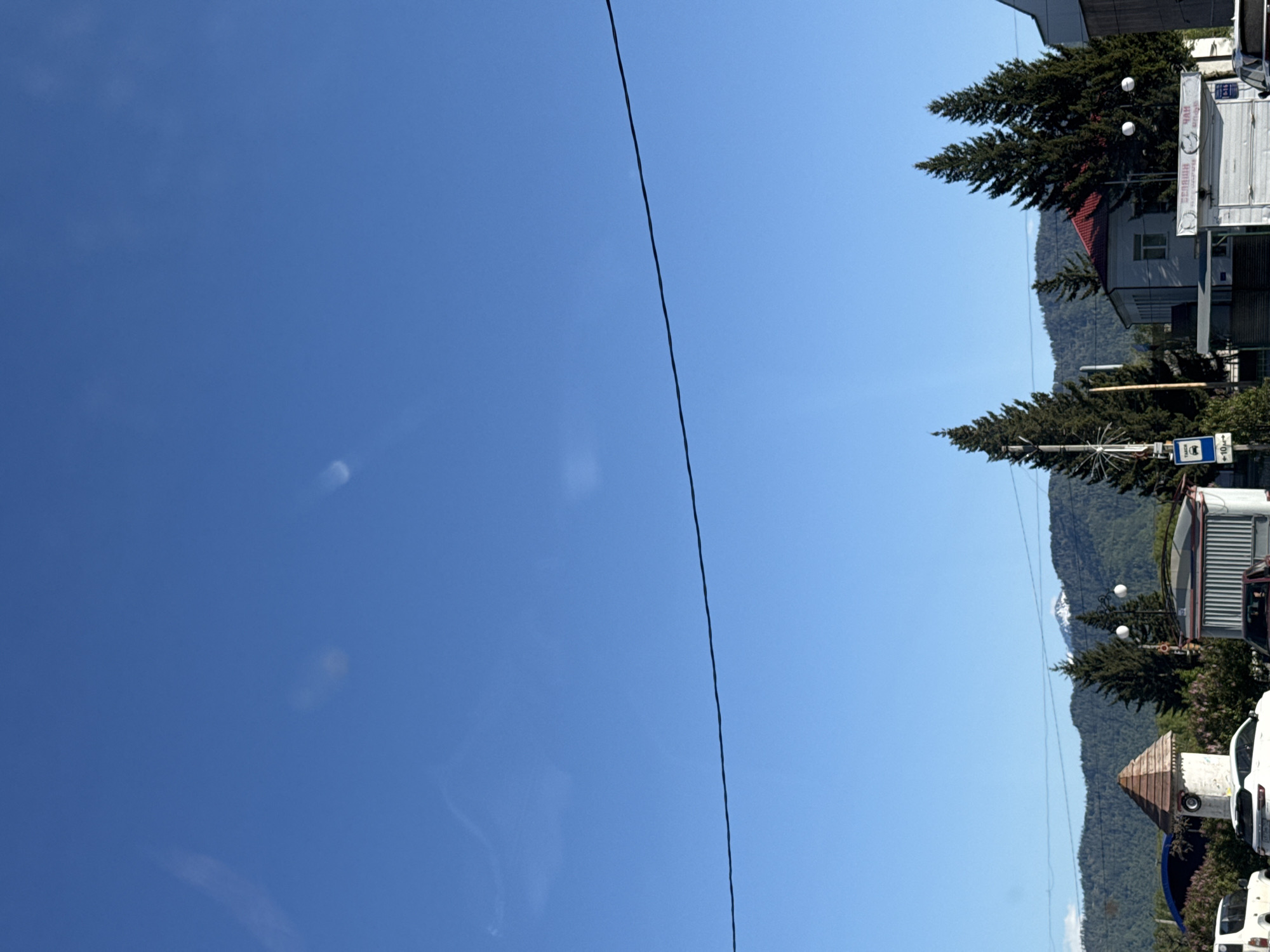The image shows a bright blue sky with a single overhead wire extending diagonally across the view. The right side of the image depicts a mountainous landscape covered in evergreen trees, interspersed with buildings. Among the buildings, there's a house with a red roof, a bus stop sign, and parked vehicles, including at least one white van. The scene appears to be in Ust-Koksa, Russia, given the geographic features and architectural elements.