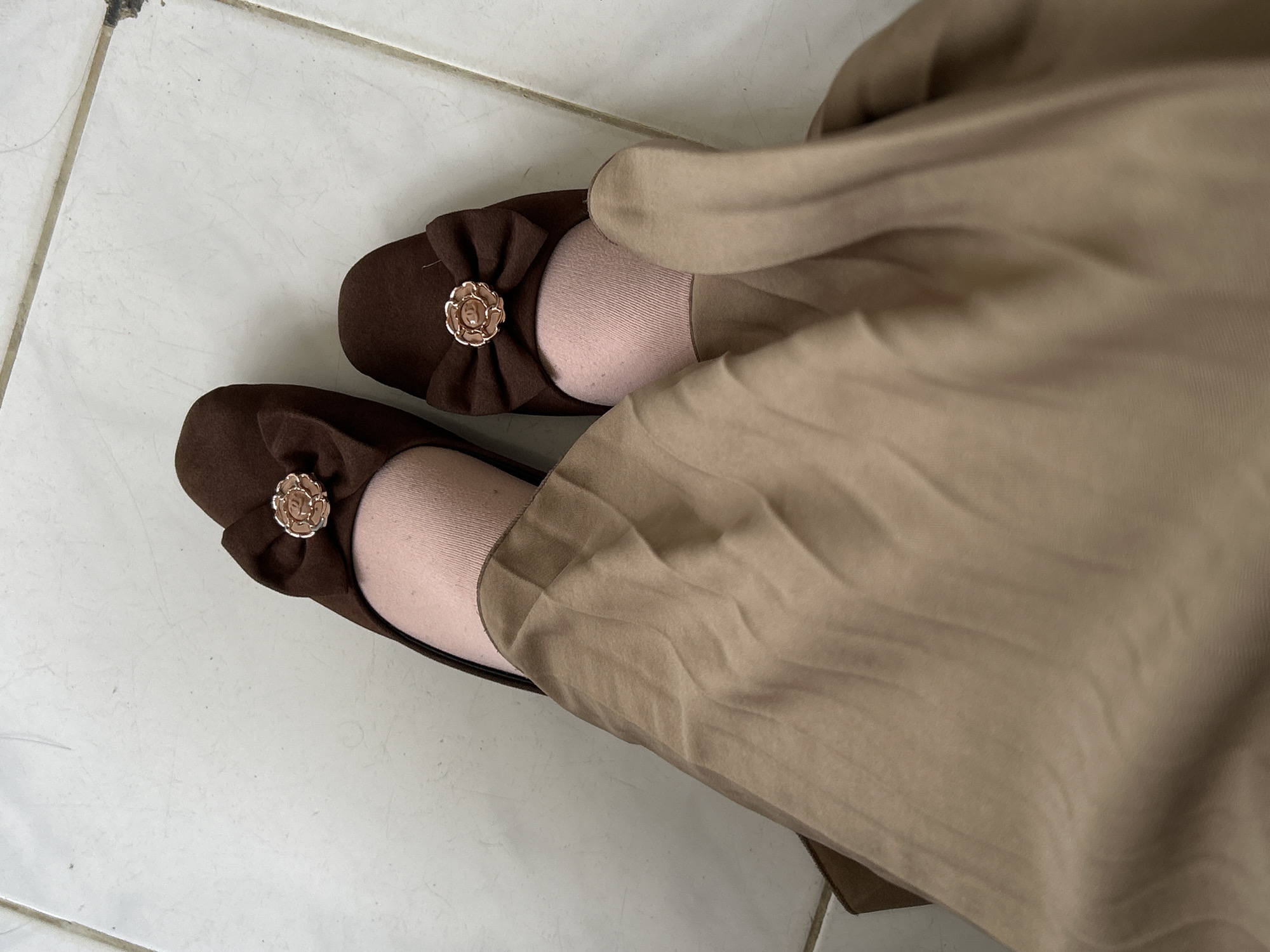The image, captured in Reuleut Barat, Indonesia, presents a top-down view of a person's feet and lower legs. The individual is wearing a pair of dark brown flat shoes, possibly made of suede or a similar soft material. Each shoe features a decorative element on the front: a small bow made of the same dark brown material, centered with a detailed, rose-shaped metallic embellishment that appears to be rose gold or antique gold.
Light pink or nude-colored socks are visible, peeking out between the shoes and the rest of the clothing. The person is wearing a long, flowing skirt or dress in a light brown, tan, or beige color, which falls over the legs, creating soft folds and drapes.
The feet are positioned on a white or light-colored tiled floor with visible dark grey grout lines, suggesting an indoor or sheltered outdoor setting. There is no discernible specific activity taking place other than standing or posing. The lighting is soft and diffused, making the time of day and external weather conditions impossible to determine. No text is visible in the image.