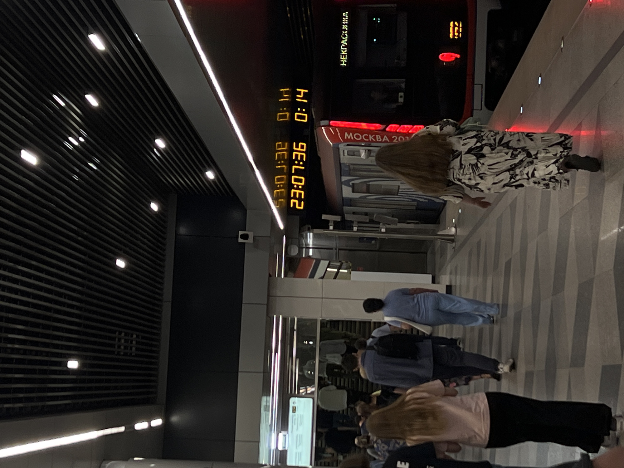 The image shows a platform in a Moscow Metro station in Russia. A train is visible on the right side, with digital signage displaying "23:07:36" and "0:14," presumably indicating time and perhaps train arrival information. The train carriage reads "MOCKBA 201" (Moscow 201). Several people are standing on the platform, some waiting near the train, while others are walking. The ceiling has a series of linear light fixtures.