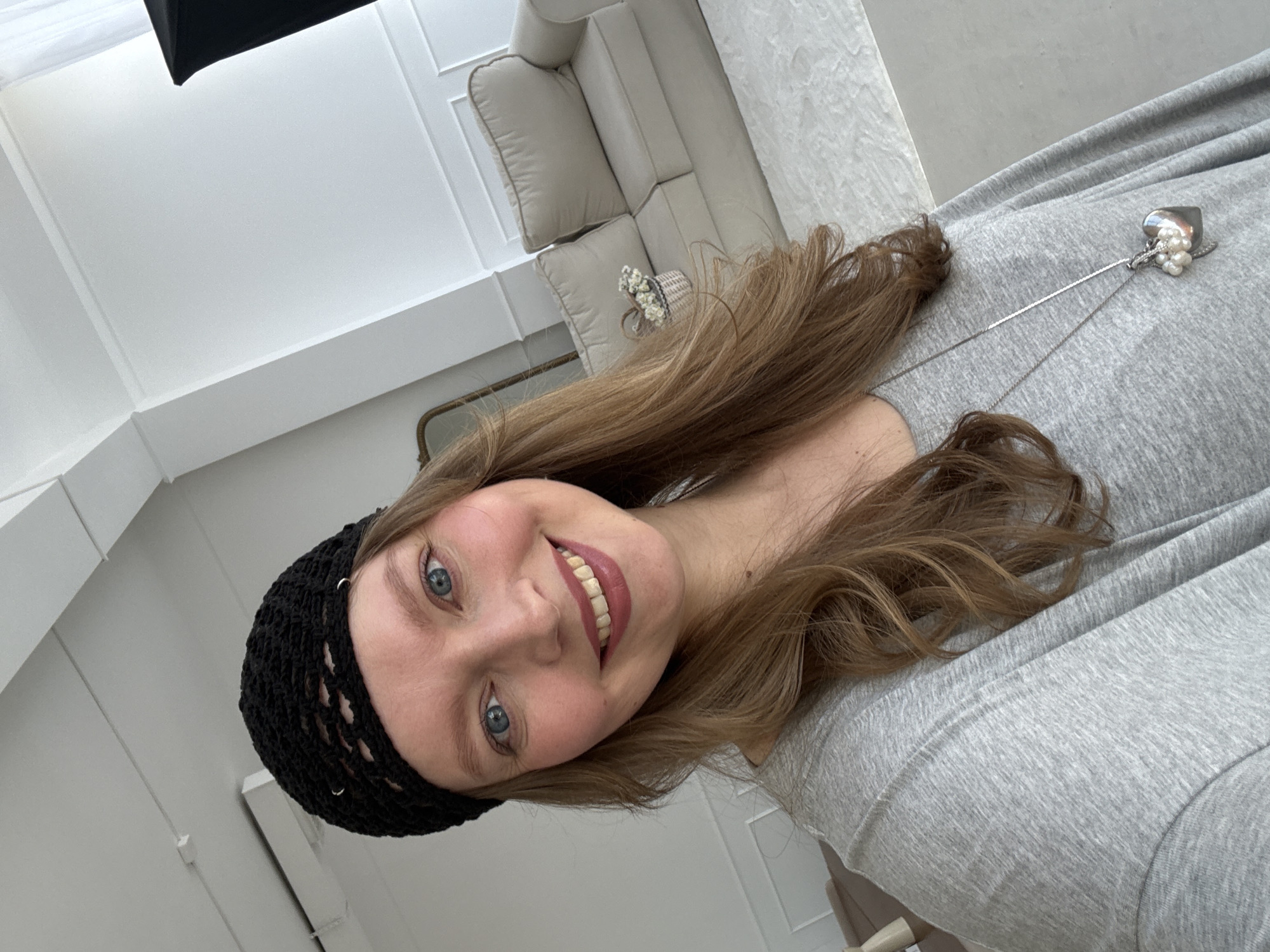 A selfie is taken by a Caucasian woman with long, light brown hair and blue eyes, wearing a black crocheted beanie, a grey long-sleeved shirt, and a silver necklace with a heart-shaped pendant. She is smiling at the camera. The background appears to be a bright, modern room with white walls, a couch, and a rug. The specific location in Izhevsk, Russia, is not identifiable from the image.