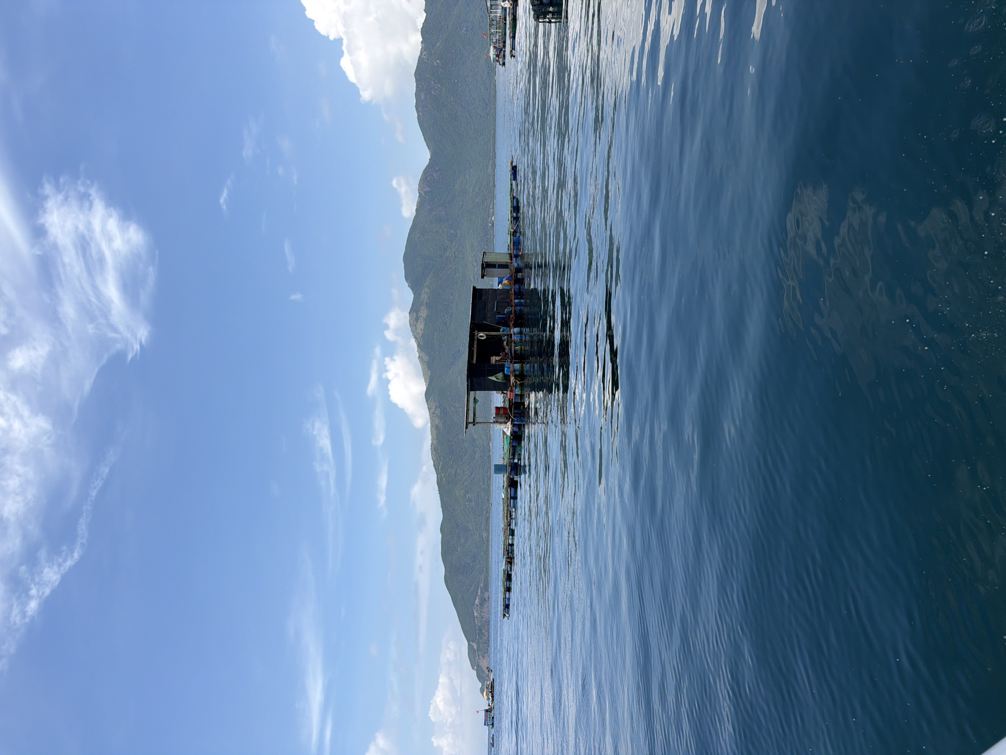 The image shows a floating structure on the sea, likely a fish farm or aquaculture setup, off the coast of Nam Nha Trang Ward, Vietnam. The water is calm and reflects the sky and surrounding landscape. Mountains are visible in the background, and the sky is partly cloudy. The structure is dark in color, possibly made of metal and wood, and appears to be several units connected together. The image is taken from water level, giving a clear view of the structure and its surroundings.