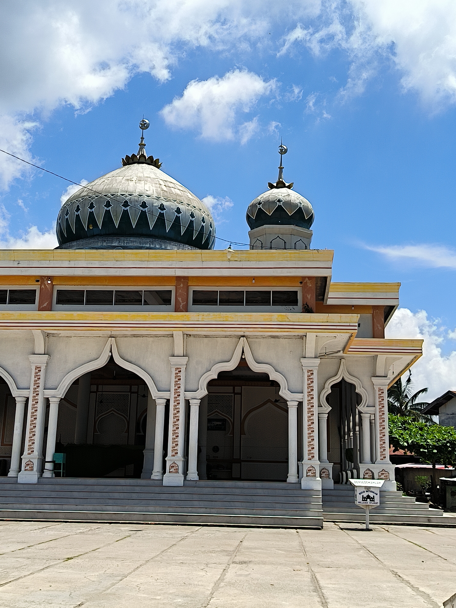 The image depicts a mosque located in Matangkruet, Indonesia. The architectural style features white pillars and arches, with decorative brickwork. Two domes top the structure, adorned with crescent moon and star symbols. The building is two stories tall. A blue sky with scattered clouds serves as the backdrop. The foreground shows a paved area leading up to the mosque, with a donation box. Greenery is visible in the background.
