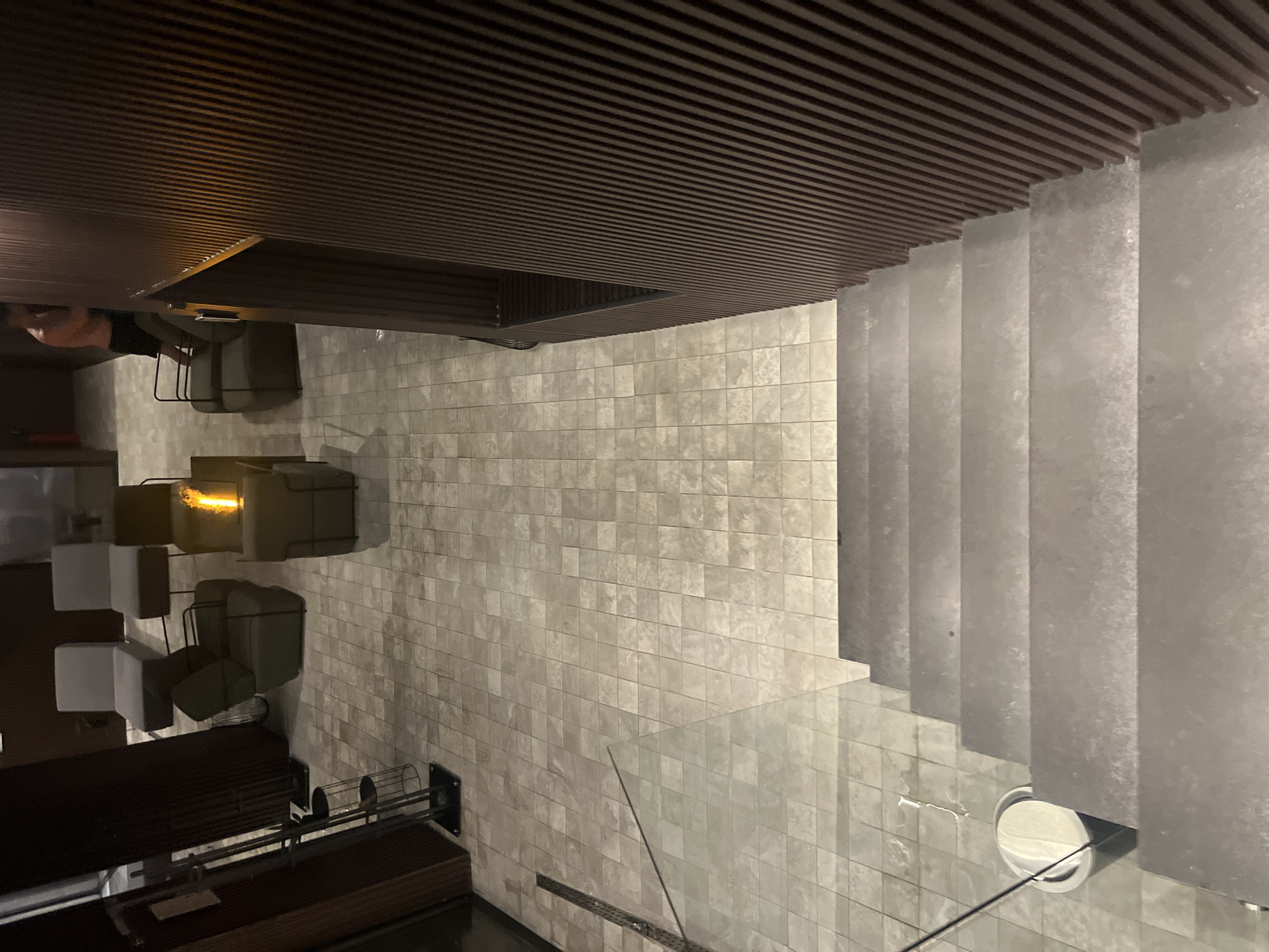 The image shows an interior with a geometrically-patterned ceiling of parallel wooden slats. To the right, concrete steps lead upwards and are bordered by a glass railing. The floor is tiled with small, light-colored tiles. On the left, there is seating including chairs and couches with a wooden table. One person is visible sitting at the table. There is a light fixture. The location may be in Sofia, Bulgaria.