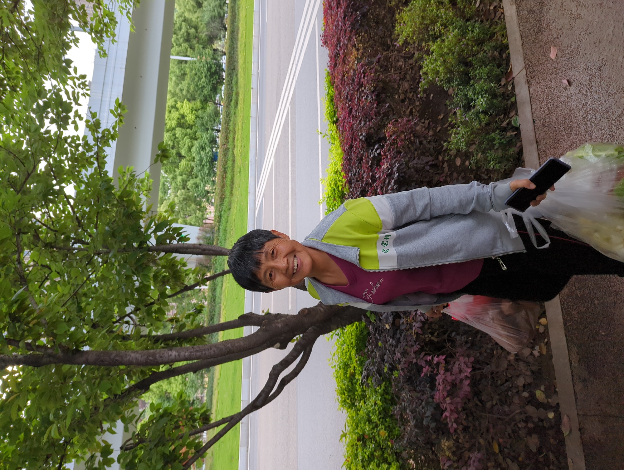 An older woman with short, dark hair smiles warmly at the camera, standing outdoors in Jiangjin, China. She wears a grey zip-up jacket with lime green panels on the shoulders and upper back, and a magenta top underneath which displays the word "Fashion" in white script. White characters, possibly "纪屯", are also visible on the lime green section of her jacket. In her right hand, she holds a black smartphone, and in her left arm, she carries a large, clear plastic bag filled with light green leafy produce. Another plastic bag is visible near her feet.
She is standing on a paved area next to a curb. Behind her, a landscaped strip features rows of bushes, including bright green foliage and reddish-purple leafy plants. Beyond the bushes, a road or paved path with two white lane markings is visible, flanked by a grassy area and distant trees. On the left side of the frame, a large tree with green leaves partially obscures a light-colored concrete structure in the background, possibly an overpass. The scene is brightly lit, indicating daytime and clear weather. The woman's broad smile conveys happiness and friendliness.
